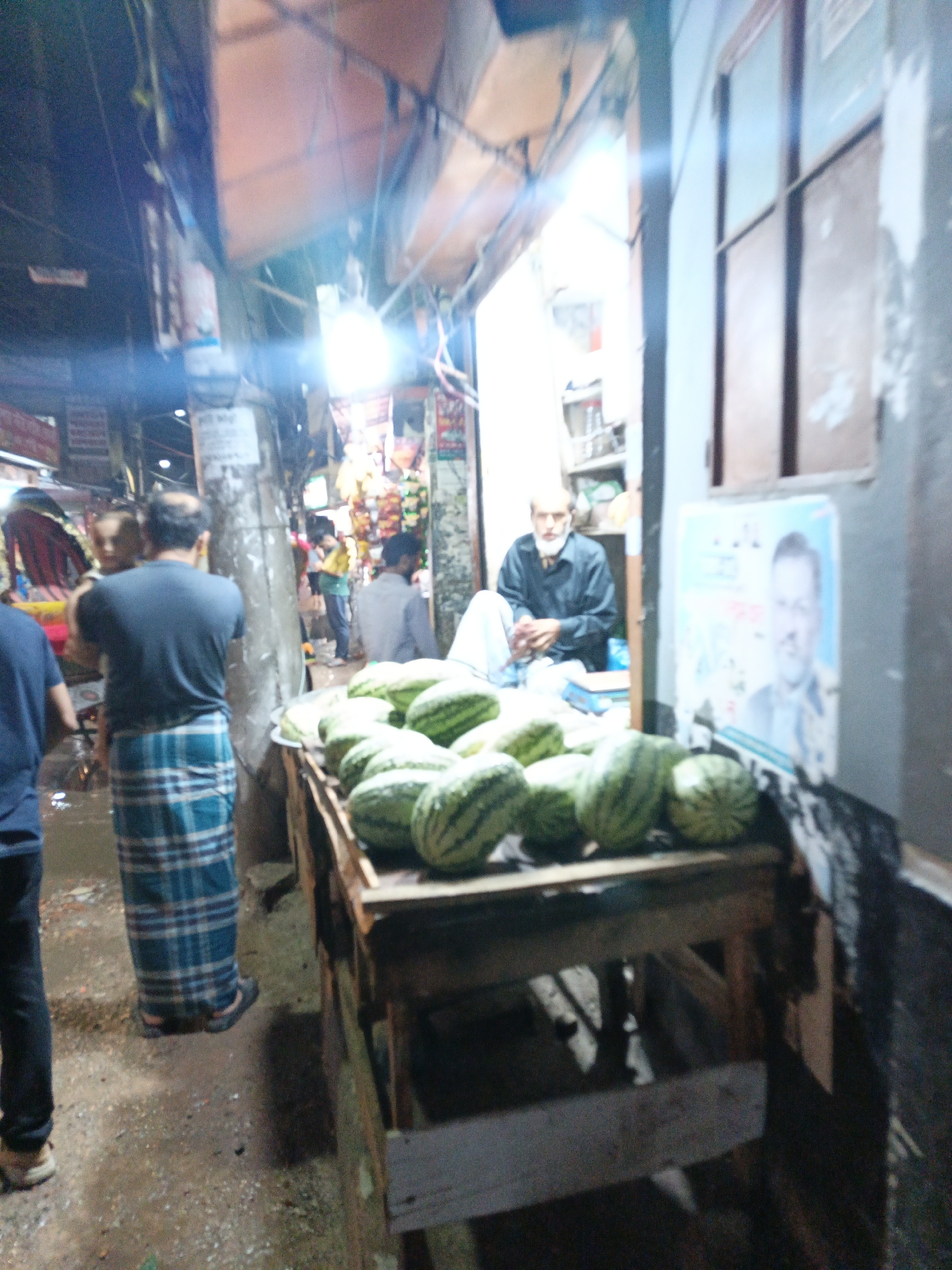 The image shows a street scene with a fruit stand selling watermelons. The location appears to be a narrow street with shops on either side. A man with a white beard sits behind the watermelon stand. There is a wooden stand displaying many watermelons in the center of the image. People are walking in the street. The overall lighting is bright, possibly due to artificial lights and daylight. The location is Unknown, Unknown.