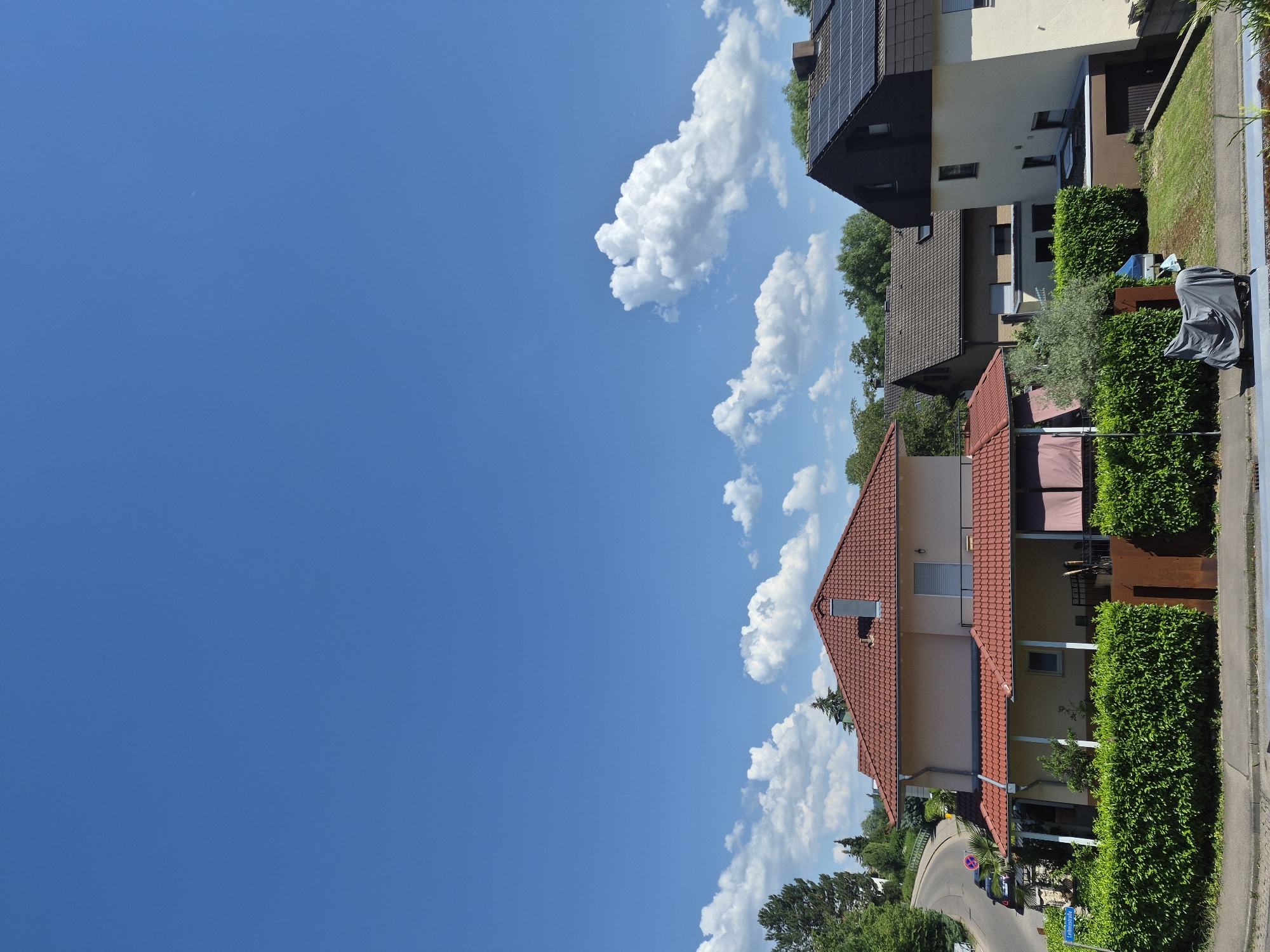 The image shows a street with several houses, a blue sky with white clouds, and green trees. The houses have red tile roofs and are surrounded by trimmed hedges. One house has solar panels on its roof. The time of day appears to be daytime, with bright sunlight casting shadows. The weather is clear with some fluffy clouds. A street is visible winding through the neighborhood, with a car parked on it. There is also some green grass and a sidewalk. In the foreground, a gray cover is draped over an object that resembles a motorcycle or a grill. One of the cropped images shows a street sign with text, but the text is illegible due to the resolution. The overall impression is a peaceful suburban scene on a sunny day.