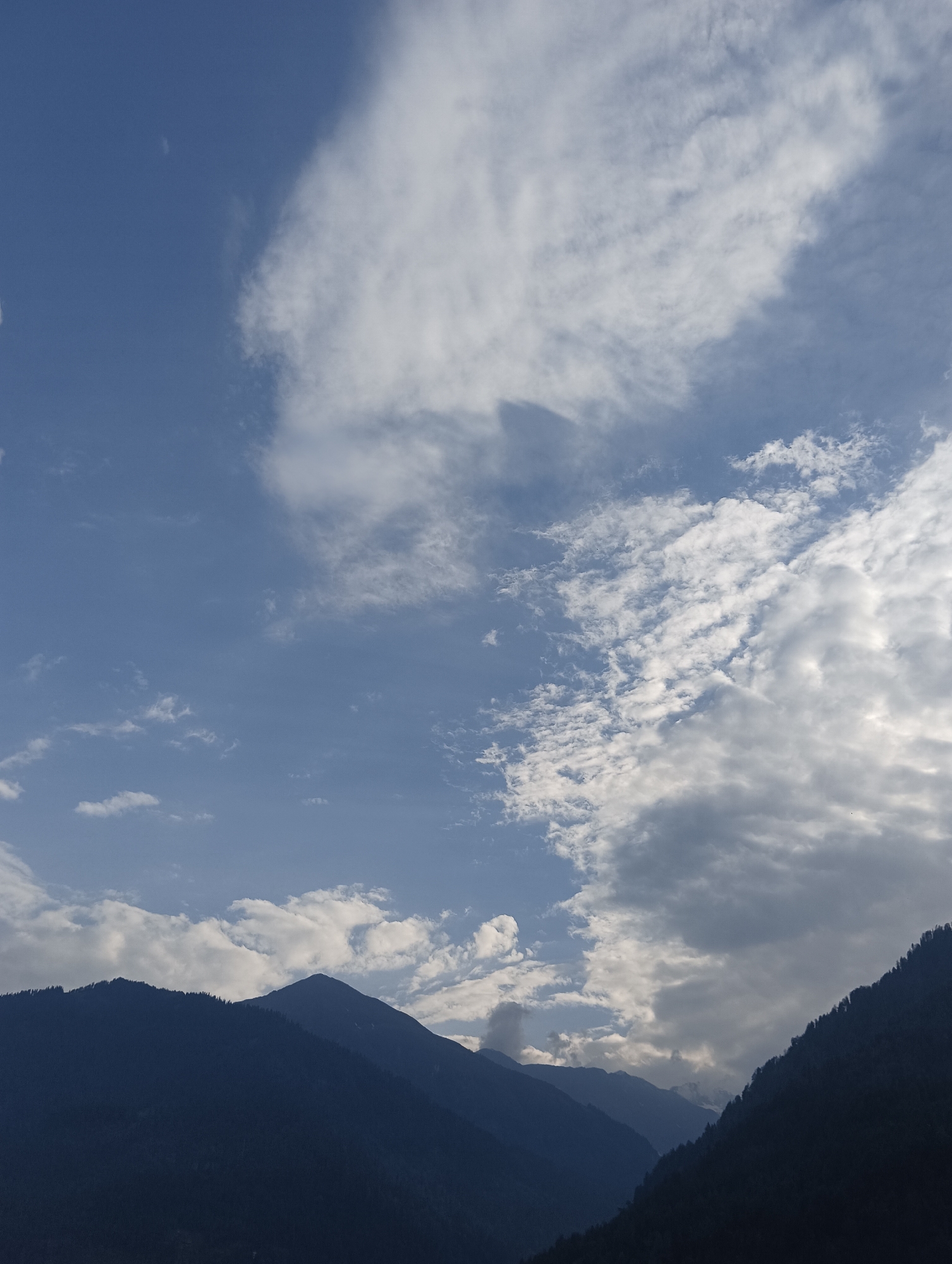 The image shows a panoramic view of mountains in Vashisht, India, against a backdrop of a blue sky filled with scattered clouds. The mountains are covered in a dark, dense forest. The sky transitions from a clear blue on the left to a more overcast and cloudy section on the right, with sunlight filtering through in some areas, creating a dynamic interplay of light and shadow on the cloud formations. The overall scene conveys a sense of natural beauty and tranquility.