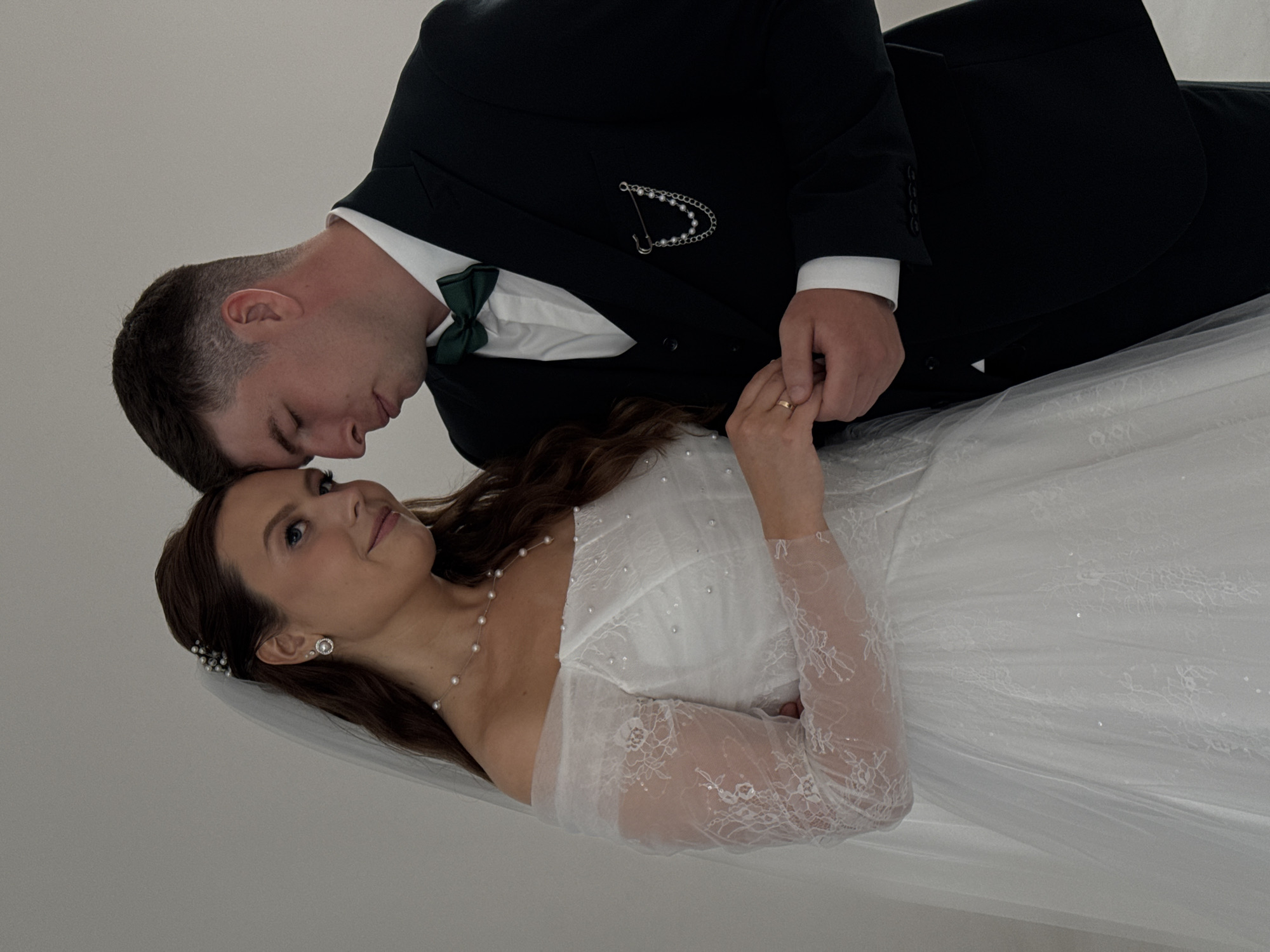 An intimate portrait captures a bride and groom, likely during their wedding in Chaikovsky, Russia. The couple is positioned against a plain, light-colored backdrop, suggesting an indoor setting for this close-up shot.
The groom, with short dark hair, is seen leaning down from the upper left side of the frame, his head tenderly close to the bride's. He is dressed in a dark suit, a crisp white shirt, and a dark green bow tie. A decorative silver pin, featuring a string of pearls, adorns his lapel.
The bride, located in the lower right, looks up at him with a soft, affectionate expression. She has long, dark hair styled with a pearl hairpiece and a delicate white veil. Her wedding gown is white, featuring a strapless bodice and elegant sheer lace sleeves. She wears pearl stud earrings and a delicate pearl necklace. Their hands are gently clasped together in the center of the image, showcasing a wedding band on the bride's finger. The overall mood is one of tenderness, affection, and marital joy. There is no visible text in the image.