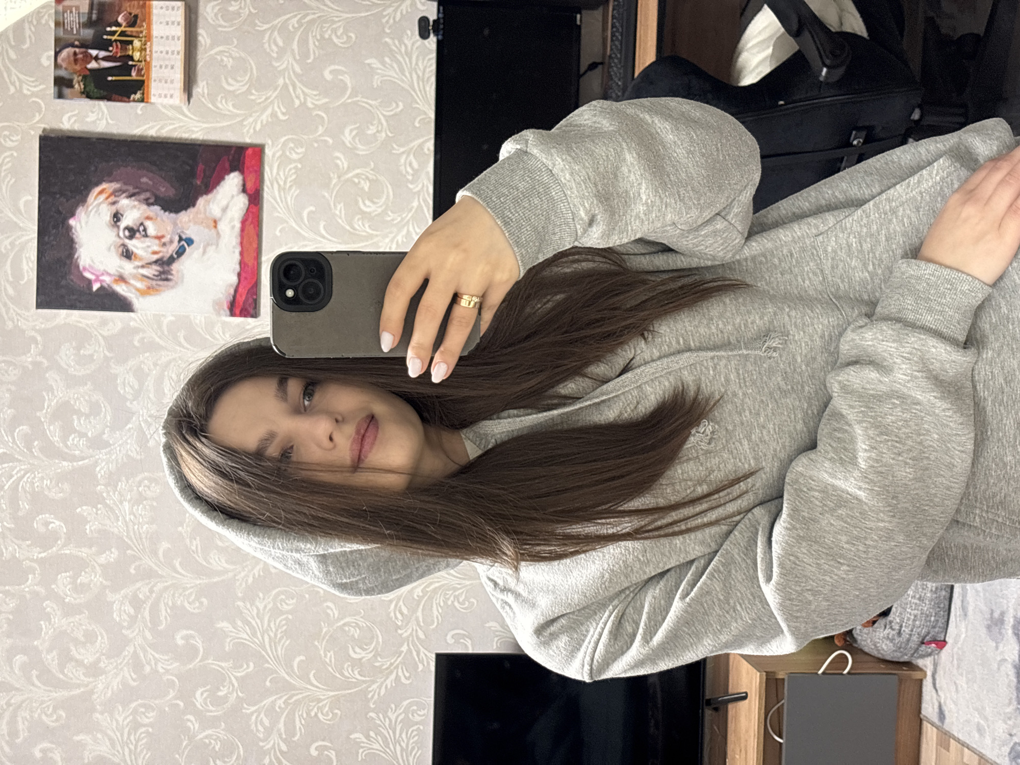 The image displays a young woman taking a selfie indoors, likely in her home in Chaikovsky, Russia. She holds a dark-colored smartphone with a prominent triple-lens camera in her right hand, which features a gold-colored ring on her ring finger and light-colored nail polish. Her face, partially visible from under the hood of her grey sweatshirt, shows a calm expression with a slight smile. Her long, dark brown hair cascades over her shoulders.
The background reveals a wall covered with light-colored wallpaper featuring a subtle, ornate floral or damask pattern. Two items are mounted on the wall: a small calendar or card depicting an older man in a suit, possibly a religious or political figure, with small, unreadable text that appears to be in Cyrillic script. Below this, a framed painting or print of a white, fluffy dog, adorned with a pink bow and a blue collar, is visible against a dark background with a red accent. Further in the background, elements of the room include a black object (possibly furniture), a dark screen (likely a TV or monitor), and a wooden surface with cables. The scene conveys a casual, personal indoor setting.