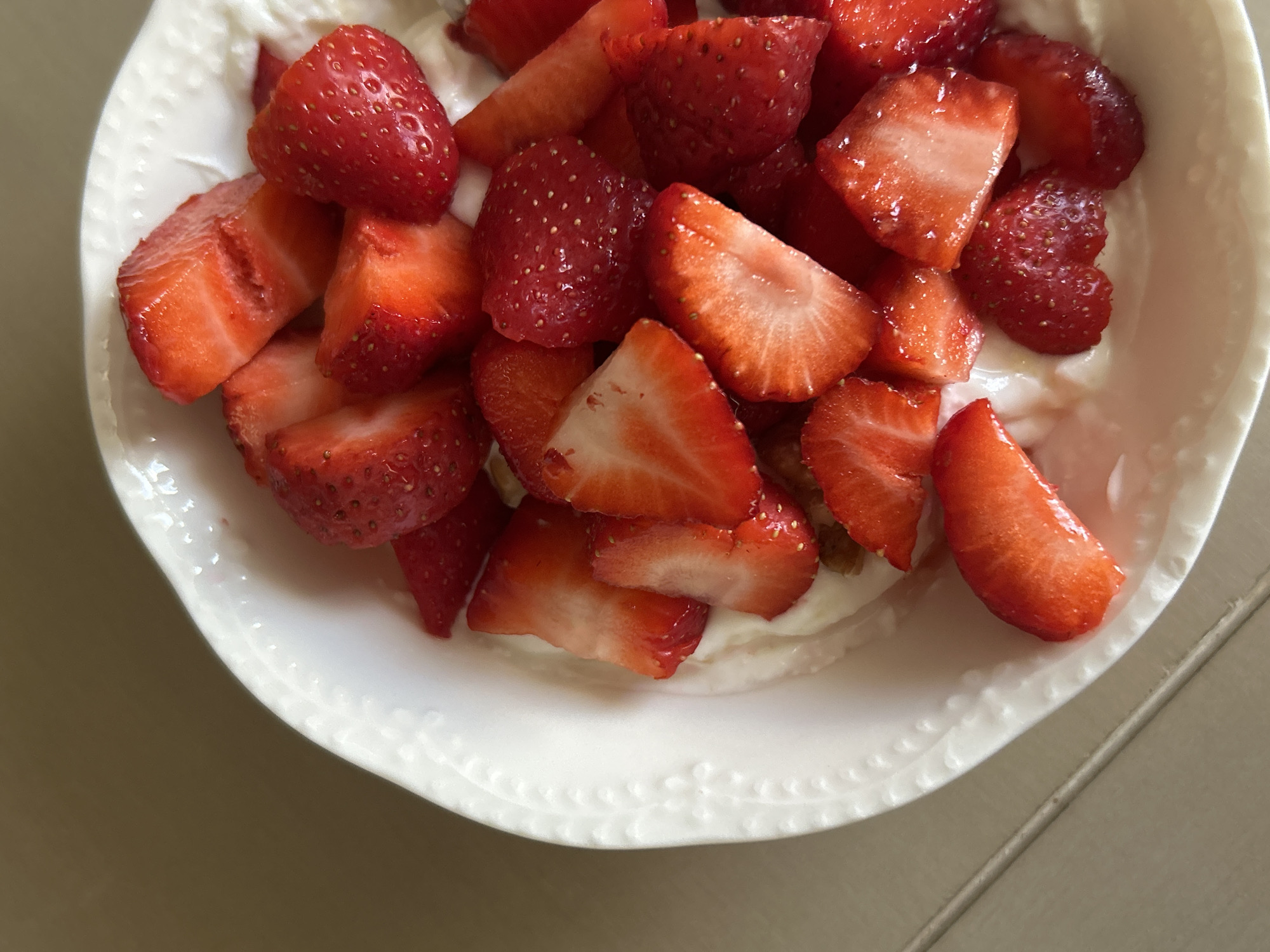 A close-up, overhead shot shows sliced strawberries on top of yogurt in a white, textured bowl. The strawberries are bright red and appear fresh, some whole and some cut in half or quarters. The yogurt is creamy and white, filling the bottom of the bowl. The bowl has a decorative, embossed edge. The background is a neutral, speckled surface, possibly a table. There is no discernible location in Plovdiv, Bulgaria.