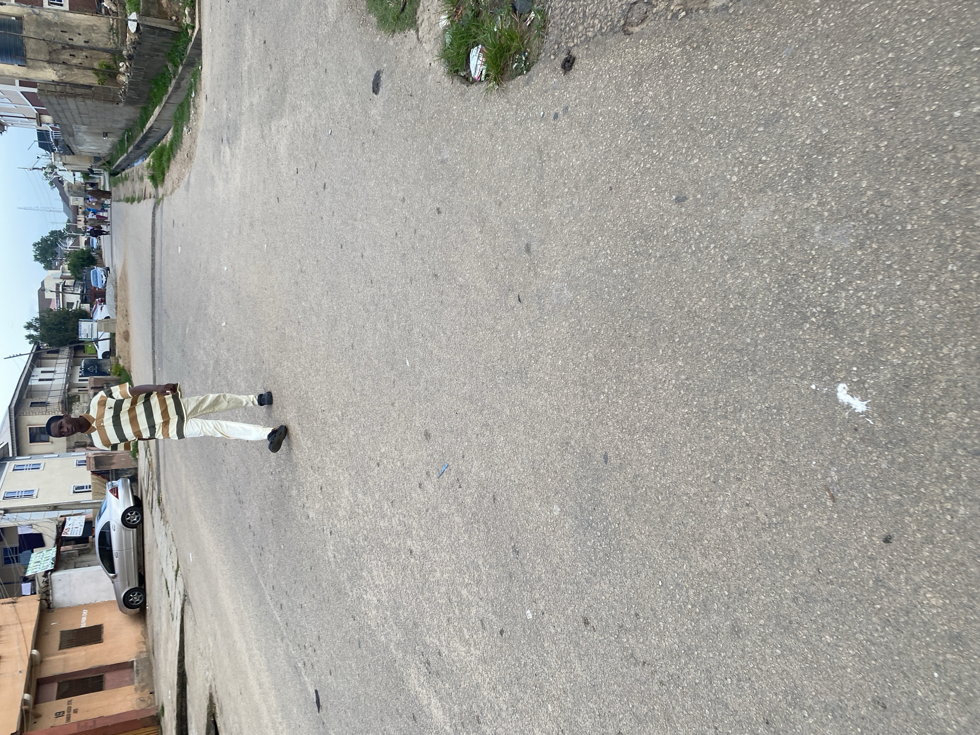 A young man wearing a horizontally striped polo shirt and white pants walks down a paved street in Jos, Nigeria. Buildings line the left side of the street, including a two-story structure with an orange facade and another with cream-colored walls. A silver sedan is parked on the left side of the road. In the distance, several people are walking away from the camera. The sky is overcast. The road surface is rough asphalt with some debris.