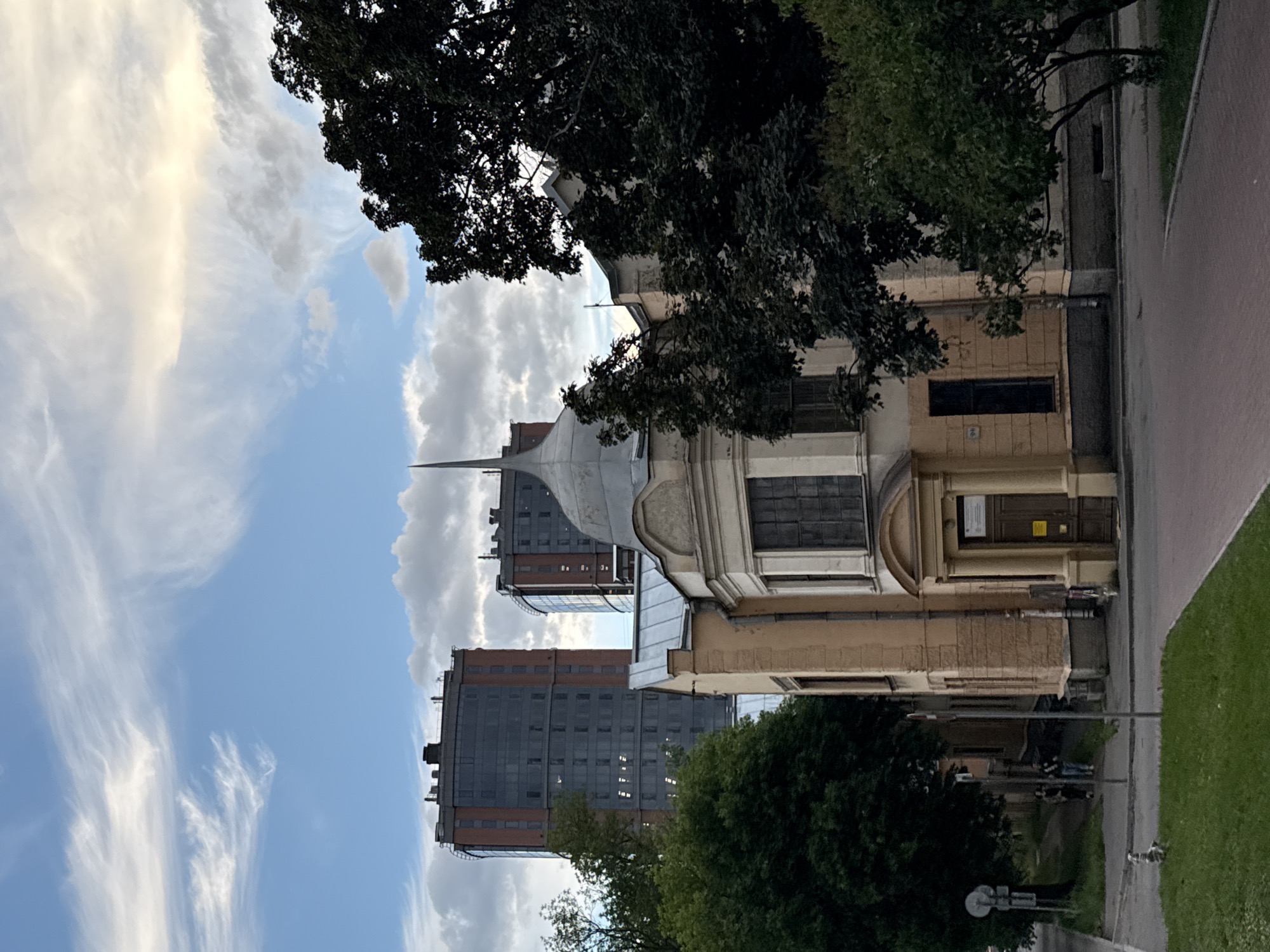 The image shows a street scene in Saint Petersburg, Russia, likely during the daytime given the bright sky. The orientation of the image is vertical, with the sky occupying the left portion and buildings and trees on the right.
The scene features a mix of old and new architecture. In the foreground, partially obscured by dark green trees, is an older, light-colored building with a distinctive domed roof and a spire. Adjacent to it, and further back, are two modern, tall buildings with dark facades, featuring numerous windows. The sky is partly cloudy, with wispy white clouds stretching across a blue expanse.
To the right of the buildings, a paved path or road runs alongside a grassy area. On the path, several people are visible, appearing to be walking or standing. One person is seated on a bench or the ground. A street lamp is also present near the path. The overall atmosphere suggests a relatively calm day in an urban environment. The lighting and cloud formations hint at it being late afternoon or early evening, but it is difficult to be precise.
No discernible text is visible in the image. The scene combines elements of nature (trees, sky, grass) with urban infrastructure and architecture, creating a layered and dynamic view of a city street.