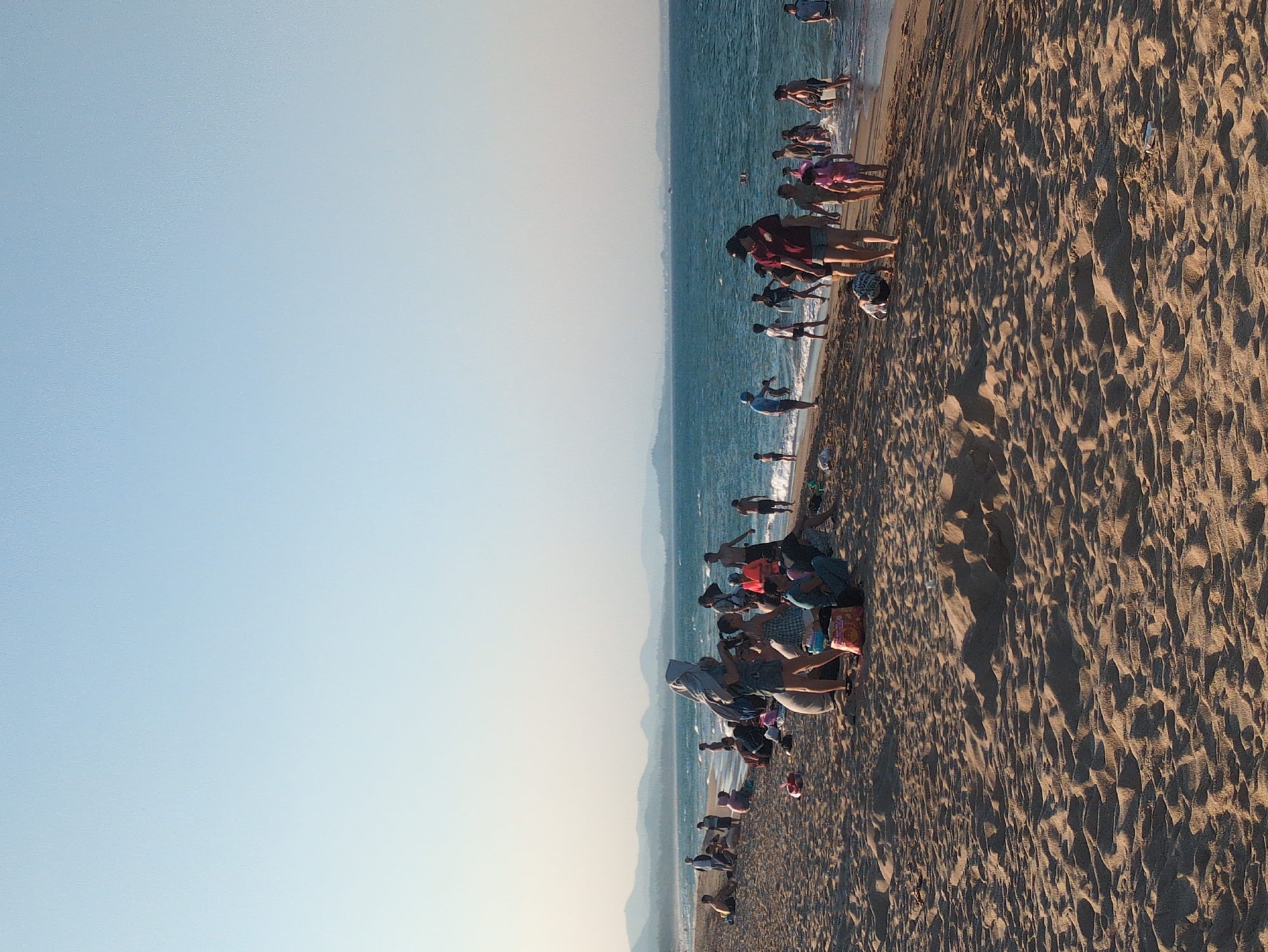 The image depicts a beach scene in Tuy Hòa, Vietnam. A stretch of sandy beach occupies the right side of the frame, with visible footprints and debris. The ocean borders the beach, with people swimming and wading in the water. A group of people is gathered on the beach, some sitting and others walking. The sky is a clear blue. Hills are visible in the background along the horizon.
