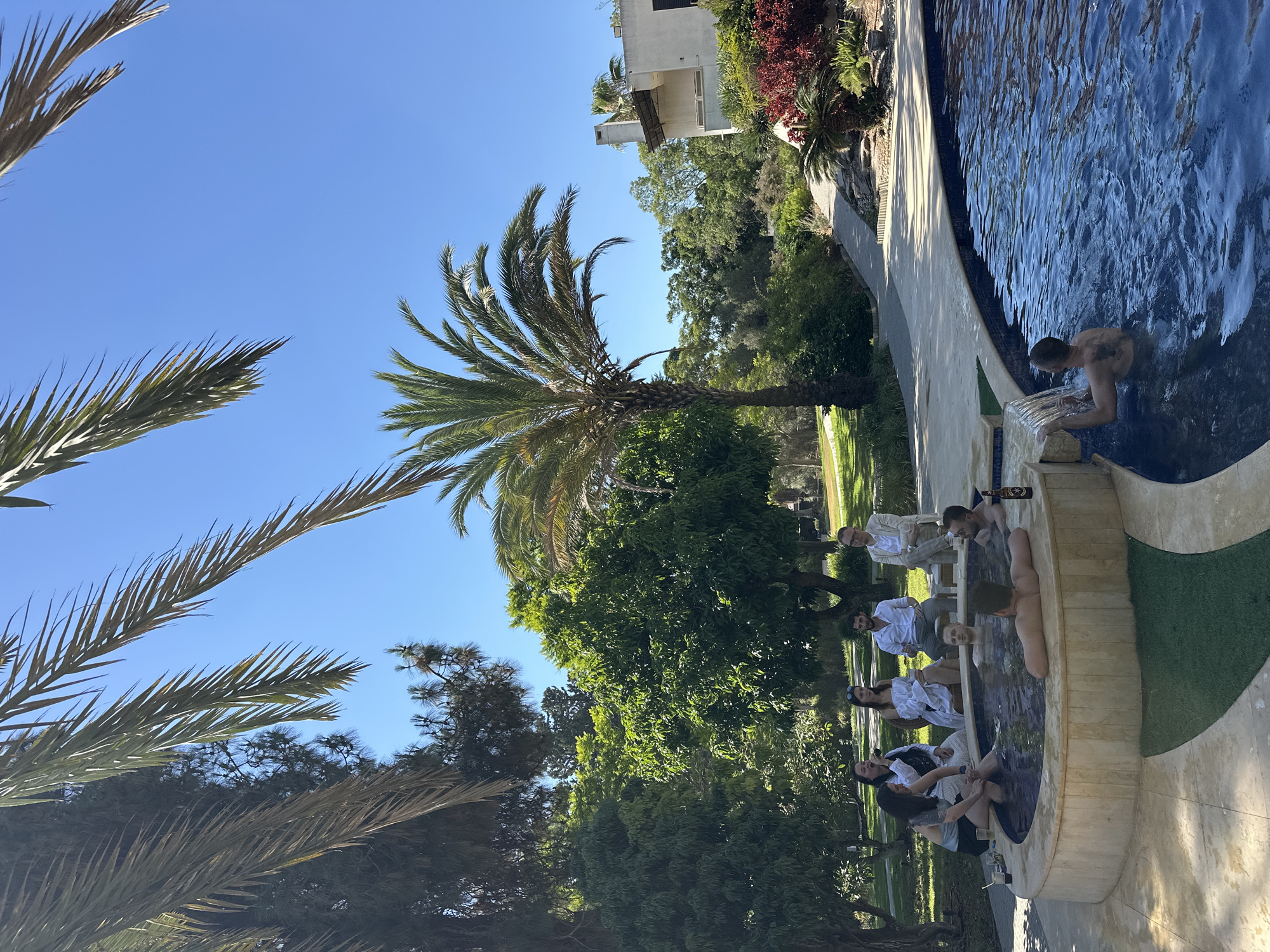 In the city of Savyon, Israel, an outdoor scene unfolds under a clear blue sky, featuring a group of people relaxing in and around a circular hot tub, adjacent to a swimming pool. The hot tub is constructed from light-colored stone and has a textured beige border. Several individuals are seated on the edge of the hot tub, some with their legs in the water, while others are submerged within it.
A man is positioned in the swimming pool, facing away from the camera, his arms resting on the edge as water cascades over the side. Another man, also in the pool, is looking towards the left of the frame. A bottle of beer sits on the edge of the hot tub.
On the left side of the scene, a group of six people are seated on the ground and on low stools. They appear to be engaged in conversation. The individuals are dressed in casual attire, with one person wearing a white shirt and another in a light-colored jacket.
The environment is lush and green, with abundant trees and foliage surrounding the pool area. Palm trees, with their characteristic fronds, are visible in the foreground and midground, contributing to a tropical ambiance. In the background, a modern building with white walls and a chimney is situated amidst the greenery. A paved walkway with stone edging leads away from the pool area. The overall impression is one of leisure and social gathering in a well-maintained outdoor setting.