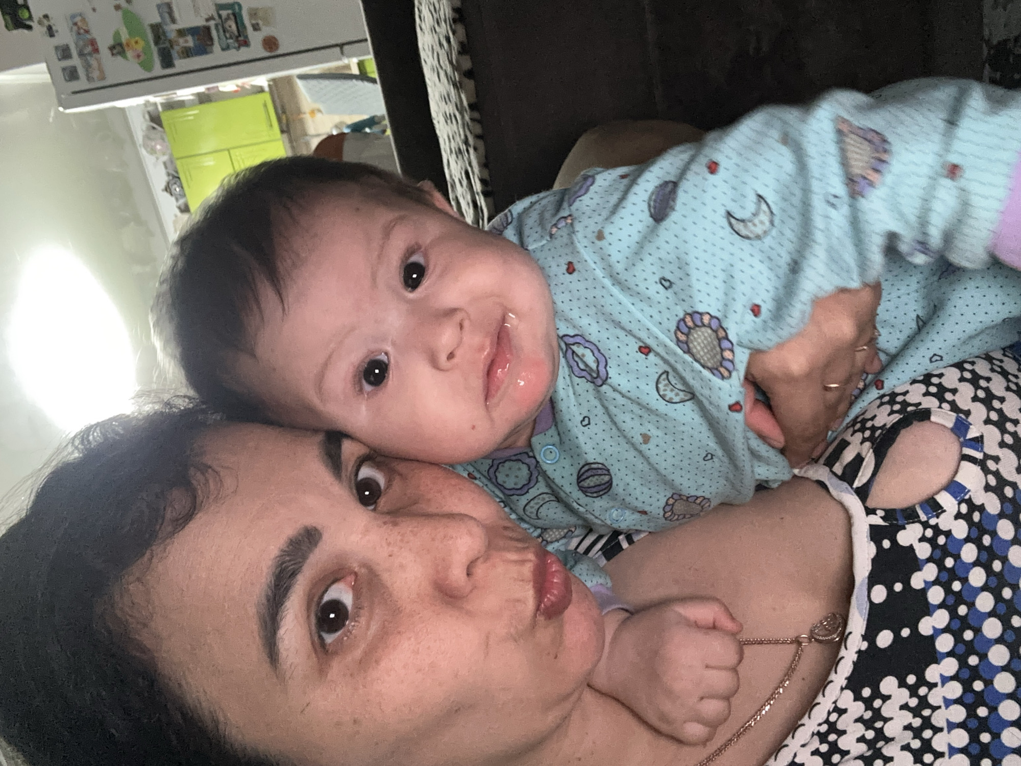 A close-up selfie captures an adult woman and a baby together indoors. The woman, positioned closer to the camera, has dark hair, dark eyes, and freckles. She is making a "pouty" or "kissing" face and wears a black, white, and blue patterned top, along with a necklace featuring a heart-shaped pendant.
Resting on or near the woman, the baby has dark hair and dark eyes. The baby wears a light blue onesie adorned with celestial patterns like moons, stars, and planets, as well as small red hearts. There is a visible smear of drool around the baby's mouth and chin. Both individuals are looking directly towards the camera.
The background suggests an indoor setting, likely a home. To the top left, a white surface, possibly a refrigerator, is visible with several small items, possibly magnets or photos, attached. A bright green object, possibly furniture or an appliance, is partially seen behind the baby's head. Darker furniture, such as a couch or chair, is visible behind the baby and to the right of the woman. No discernible text is visible in the image. The mood appears playful and intimate.