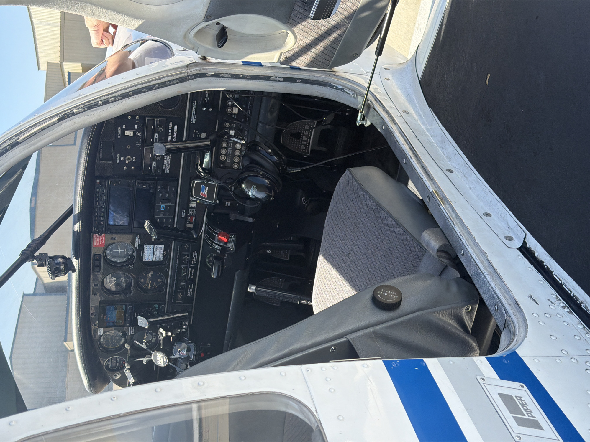 The image shows the cockpit of a Piper aircraft, featuring various control panels, gauges, and a grey cloth seat. Visible markings include "PIPER" on the aircraft's exterior and within the cockpit, as well as labels such as "SEAT RACK RELEASE," "NAV LIGHTS," and "STROBE FLASHERS." A warning sign about seat belts and no smoking is also present. The presence of a person is indicated by a hand visible near the top of the image, suggesting someone is either entering or exiting the aircraft. The context is ambiguous, but the overall setting implies an airport or aviation environment. The location in the city of Los Angeles, United States of America cannot be determined from the image.