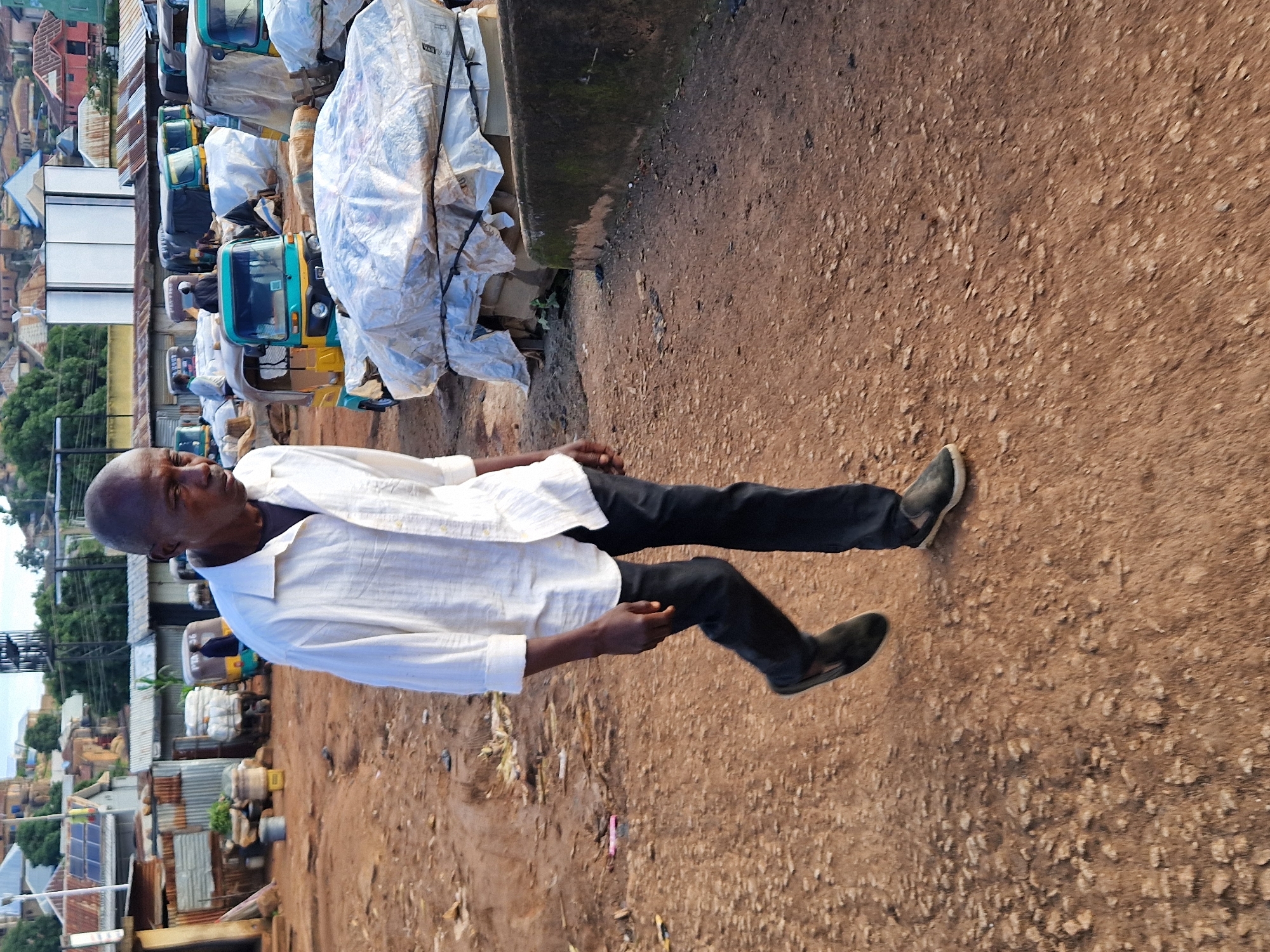 In New Stadium, Nigeria, a man walks through a dusty marketplace, his white shirt and dark trousers contrasting with the earth-toned surroundings. He is walking towards the right side of the frame, his gaze directed forward. In the background, several auto-rickshaws, some covered in plastic sheeting, are parked. Buildings with corrugated metal roofs and solar panels are visible further in the distance, suggesting a mix of residential and commercial structures. The ground is uneven and appears to be dirt or gravel, littered with some debris. The overall scene depicts a typical bustling marketplace environment in a Nigerian city.