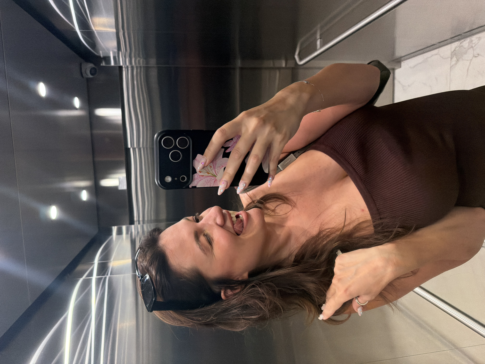 The image shows a woman taking a selfie in an elevator. She is wearing a brown top and sunglasses. Her hair is long and brown, and she has long nails with glitter. The elevator walls are made of stainless steel, and there are lights and a security camera visible. The floor of the elevator is also visible, with a marble-like pattern. The woman appears to be in a good mood, with a smile on her face and her tongue sticking out slightly. The image is likely taken during the day, as indicated by the lighting in the elevator. The location is identified as Belgrade, Serbia, but there are no specific visual cues in the image to confirm this. There is no visible text in the image.