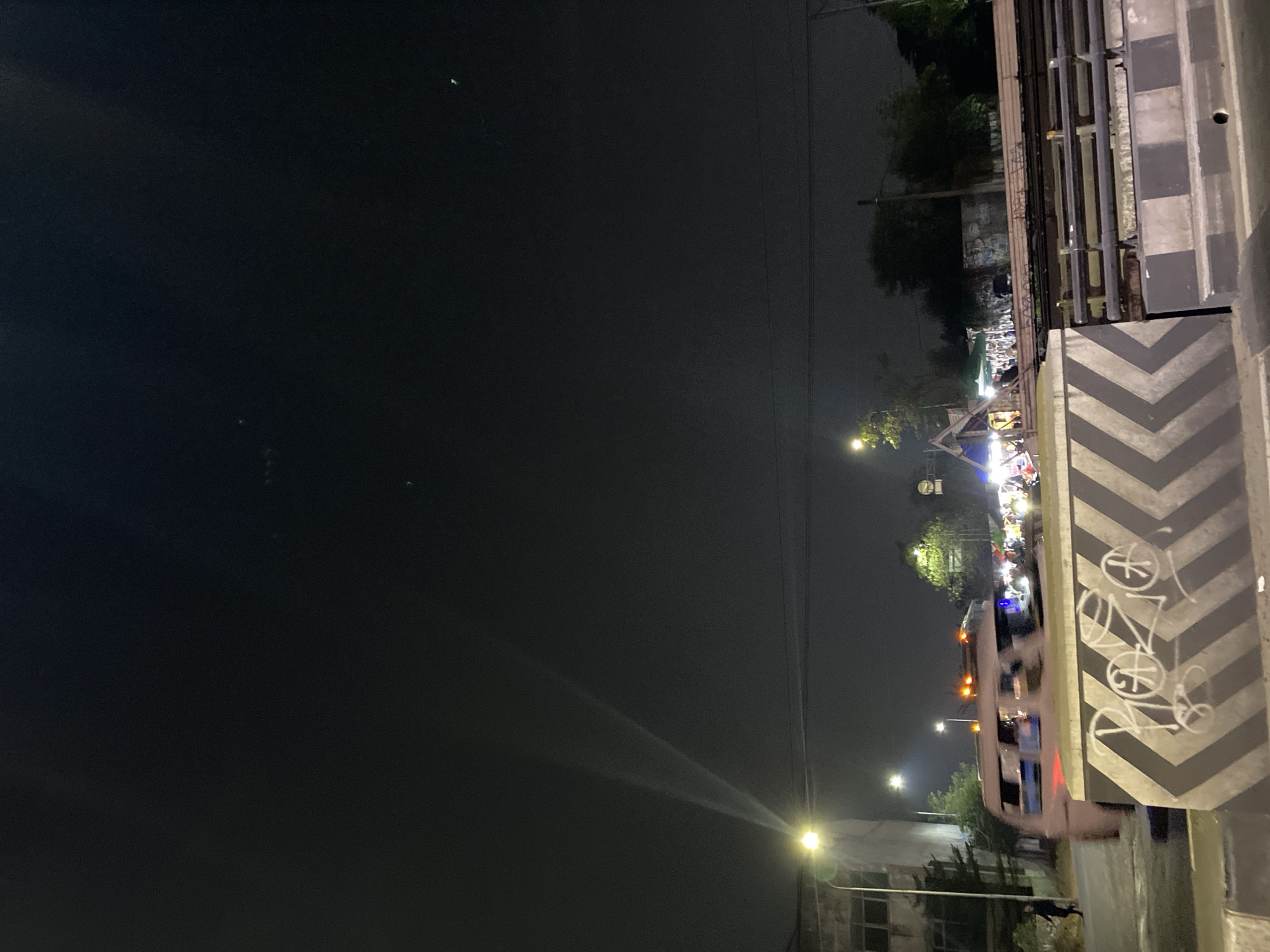 The image depicts a nighttime scene in Tangerang, Indonesia, likely a street market or roadside area. A light pole illuminates the foreground, casting two distinct beams of light upwards into the dark, hazy sky. To the left, a portion of a building with windows is visible, and a lone figure walks on the sidewalk. A light-colored van with orange hazard lights is in motion, partially obscuring a row of market stalls in the background. These stalls are illuminated by various lights, and several people can be seen browsing or working. A concrete barrier with black and white chevron markings and graffiti runs across the bottom of the frame, with metal railings visible further back, suggesting the presence of an overpass or elevated structure. Trees and other vegetation are sparsely scattered throughout the scene. The overall atmosphere is dark and somewhat murky, with artificial lights cutting through the night.