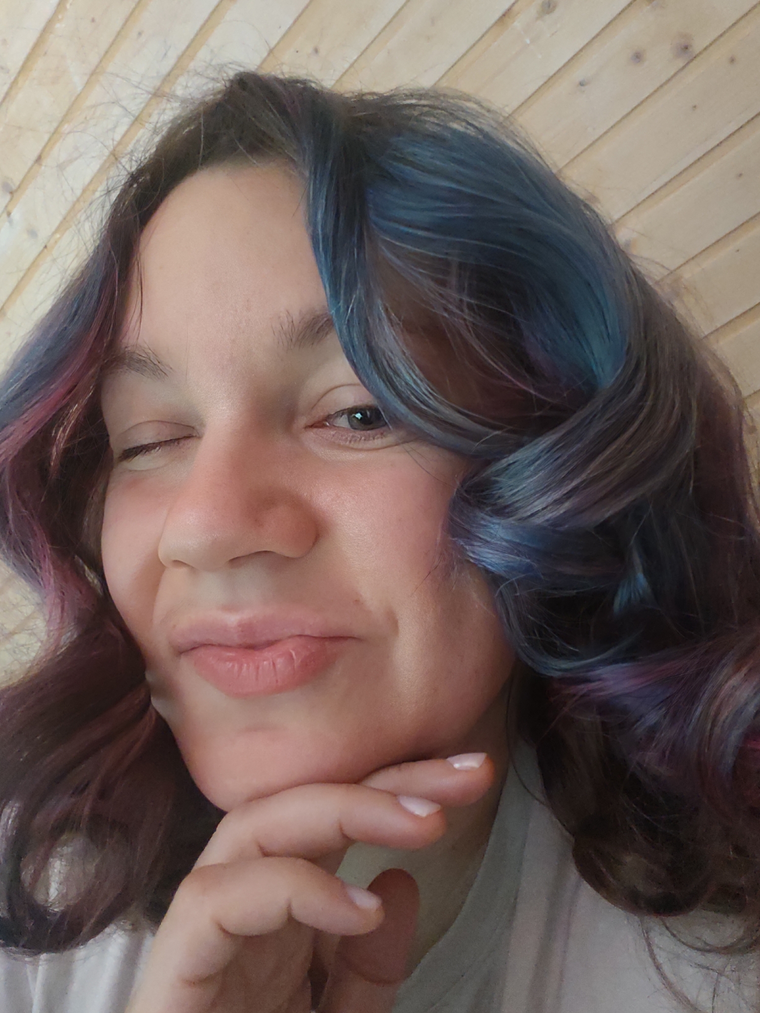 A young woman with multi-colored hair (blue, purple, and brown hues) is winking and smiling at the camera. Her left eye is closed, and her right eye is open. Her right hand is placed under her chin. The background appears to be a wooden wall. The location is not discernible from the image, and the description of Свистуха (кузяевский С/о), Russia, is not applicable.