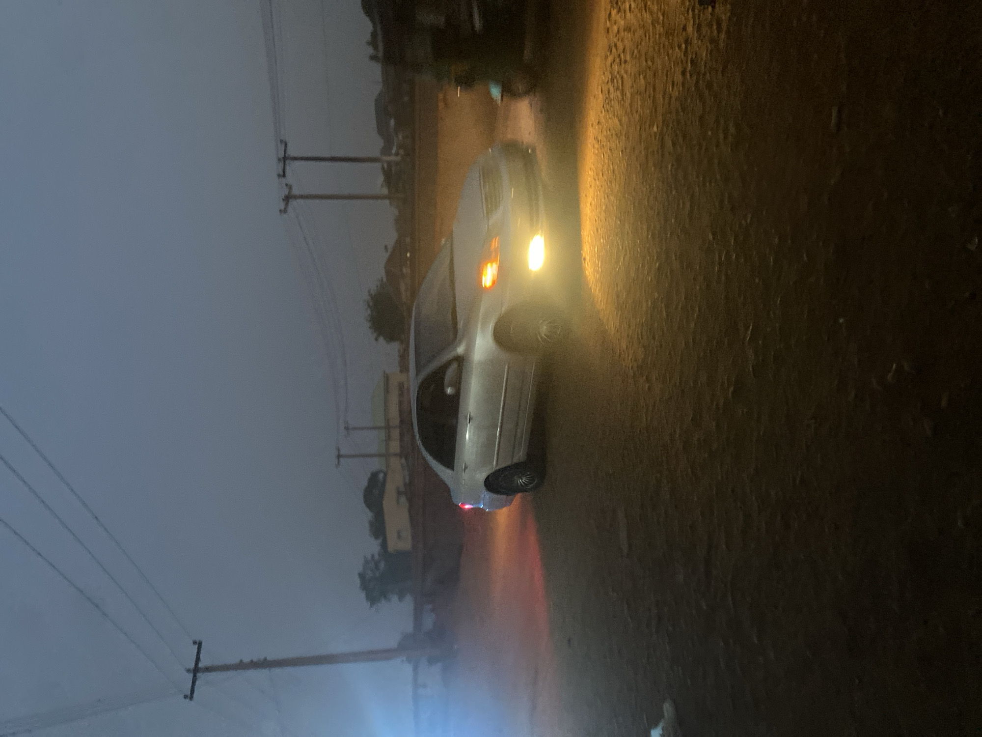 A car is parked on a wet street in Tudun Wada, Nigeria. The car's headlights and taillights are illuminated. Power lines and utility poles are visible along the side of the road. The sky is overcast. The road surface is rough.