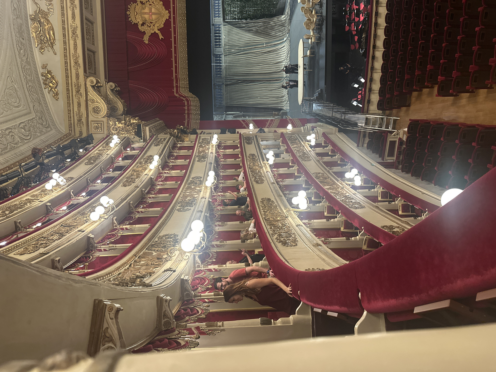 An interior view of a grand opera house shows multiple levels of ornate balconies with red velvet seating and gilded trim. Several individuals are visible on one of the upper balconies, some standing and conversing, while others are seated. The stage area is visible at the far end, with elaborate curtains and set pieces, and a few figures are present on the stage. The lighting is subdued, with numerous decorative lamps illuminating the interior. The overall atmosphere suggests a performance or rehearsal is about to begin or has recently concluded.