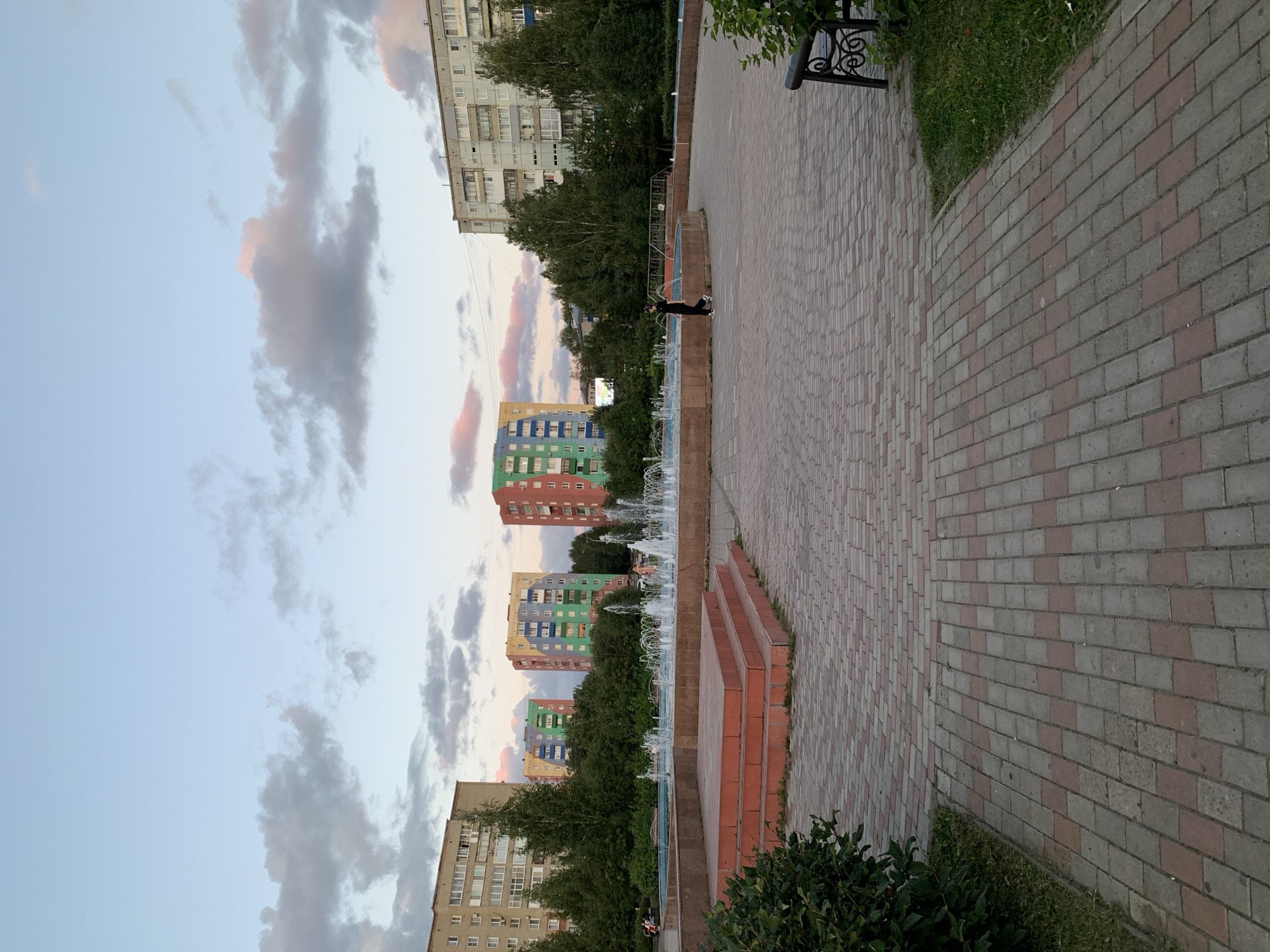 The image depicts a public square in Prokopyevsk, Russia, featuring a fountain at its center. The square is paved with interlocking bricks in a pattern of red and grey. To the left of the fountain, there are tiered brick steps. Surrounding the fountain is a circular pool with water jets active. In the background, several residential apartment buildings of varying colors are visible, interspersed with green trees. The sky above is a pale blue with scattered clouds, some tinged with pink, suggesting it might be late afternoon or early evening. A single person, dressed in dark clothing, is walking on the paved area to the right of the fountain. A park bench is partially visible on the far right of the image, adjacent to a patch of grass and bushes.