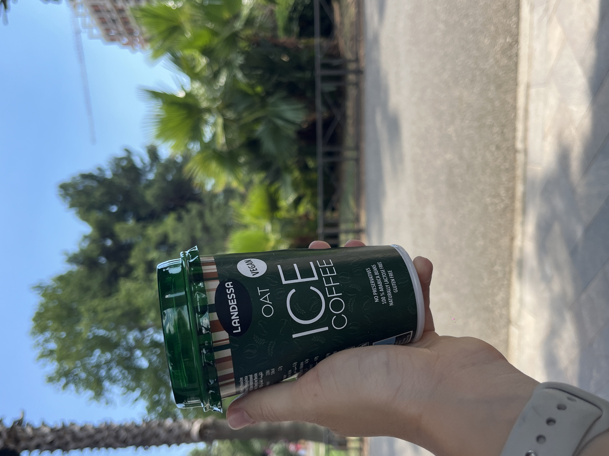 A person holds a green can of Landessa Oat Ice Coffee outdoors on a sunny day. The can has a white "VEGAN" sticker and the text "OAT ICE COFFEE" prominently displayed. Smaller text on the can indicates "NO PRESERVATIVES", "100% ARABICA BEANS", "NATURALLY LACTOSE FREE", and "GLUTEN FREE". The background features lush green trees, palm fronds, and a bright blue sky, suggesting a warm, tropical or Mediterranean setting. A blurred building and construction crane are visible in the distance, along with a paved walkway and shadows on the ground, indicating an urban or park-like environment. The lighting and shadows suggest it is daytime. The person's hand is visible holding the can, with a smartwatch on their wrist. The overall scene evokes a sense of refreshment and outdoor enjoyment.