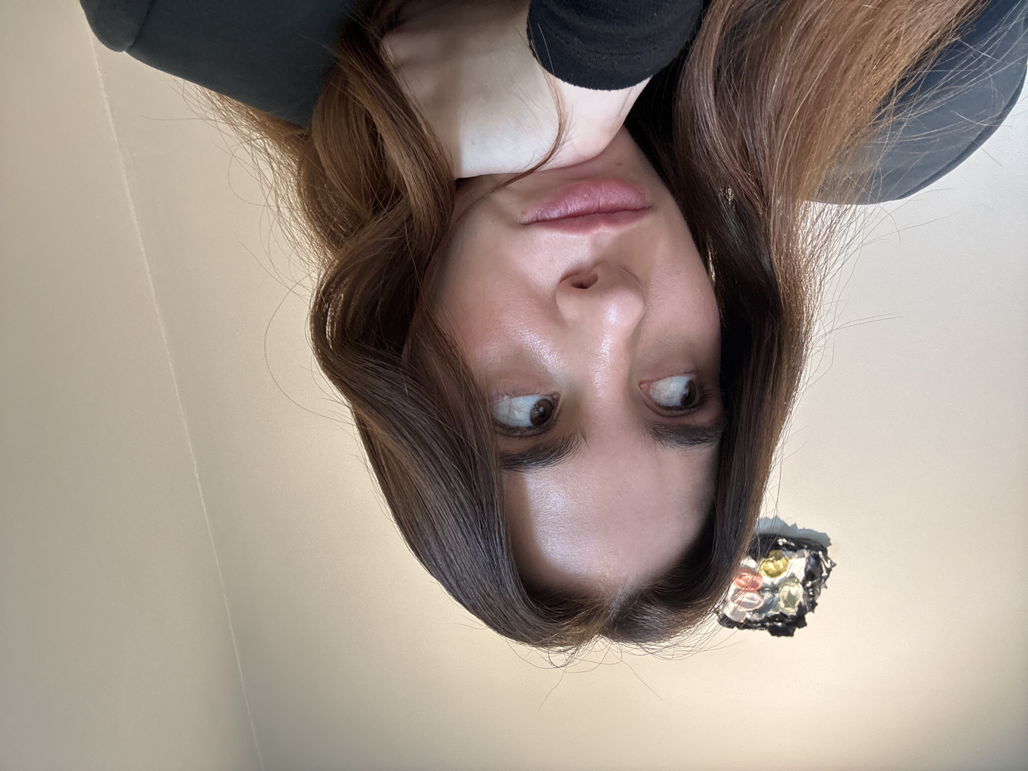 A selfie taken by a woman from a low angle, looking up. She has long brown hair, fair skin, and is wearing a dark colored top. She is resting her chin on her hand. An ornate light fixture is visible in the lower right corner of the frame. The background appears to be a plain, light-colored ceiling. The image does not provide enough information to discern a location in Paris, France.