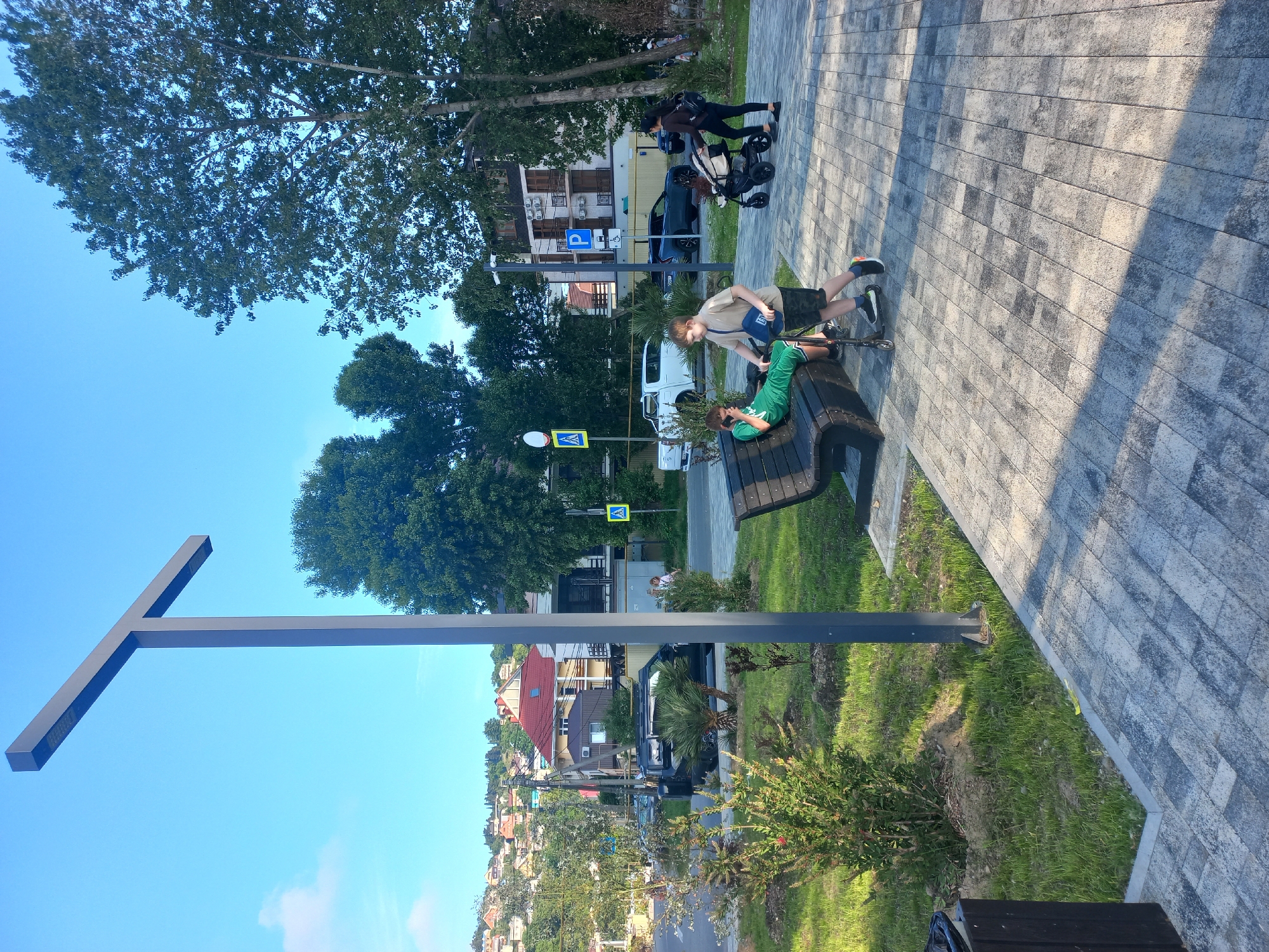 The image shows a horizontal view of a park or public space in an unidentified city, possibly a resort town. Two boys are on a park bench. One boy, wearing a green shirt, reclines lengthwise on the bench, while the other, wearing a khaki shirt and dark shorts, stands beside the bench, his posture suggesting he is either speaking to or looking after the boy on the bench. There is a black scooter to the right of the bench.
The park area is paved with gray brick. Beyond the park bench, there are trees, parking signs and pedestrian crosswalk signs. A family pushing a stroller is in the background. Across the road, a hillside is visible, covered with houses and buildings. The sky is clear and blue. A tall, black T-shaped lamppost is in the foreground.