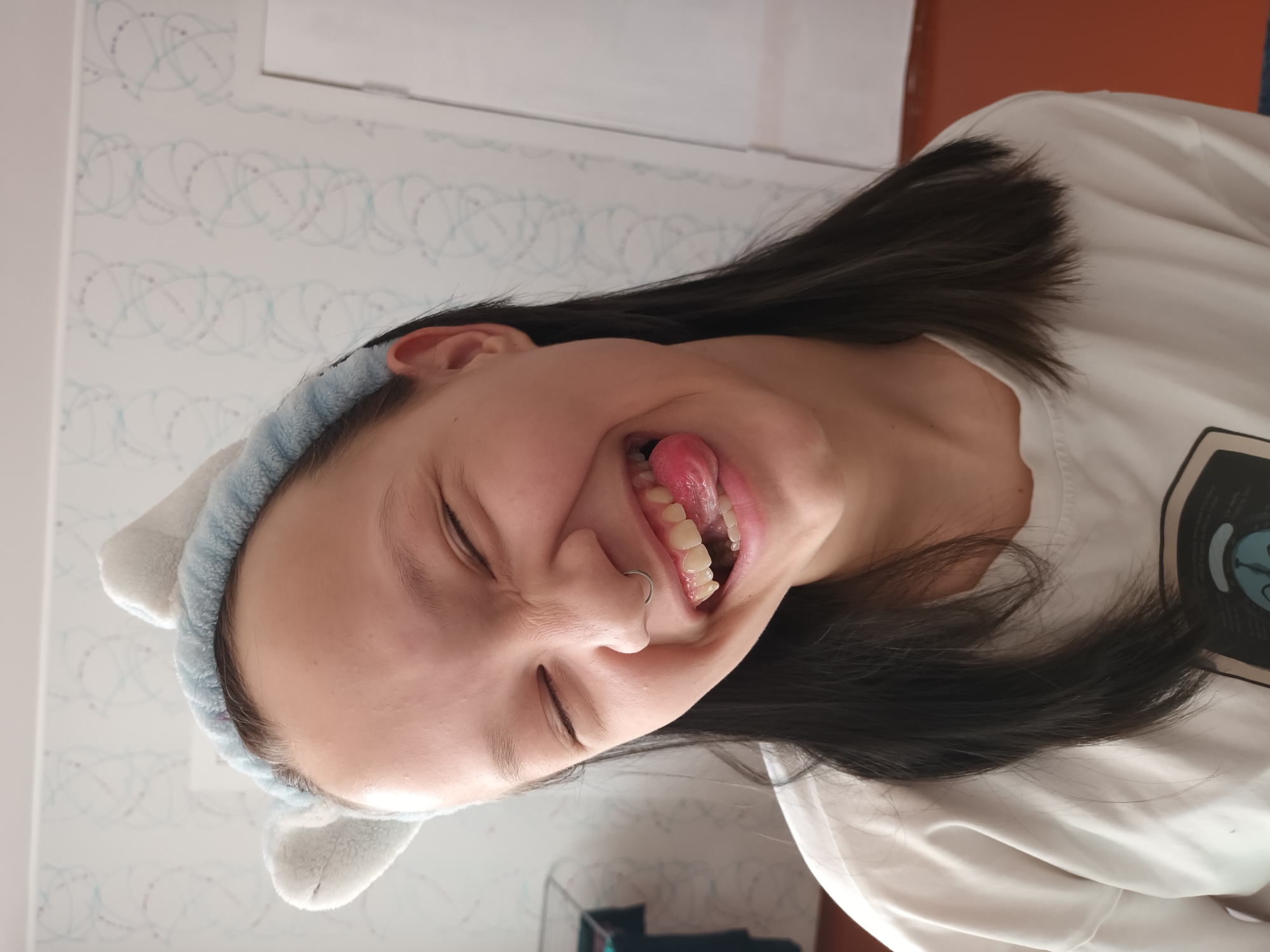A young woman is indoors, positioned sideways with her head tilted to the right. She is wearing a light blue headband with white cat ears. Her eyes are closed, and she is sticking her tongue out. She has dark hair and a nose ring. She is wearing a white top with a black design on it. The background consists of a white wall with a light blue patterned design. It is not possible to ascertain the exact location in the city of Ольховка, Russia, from this image.