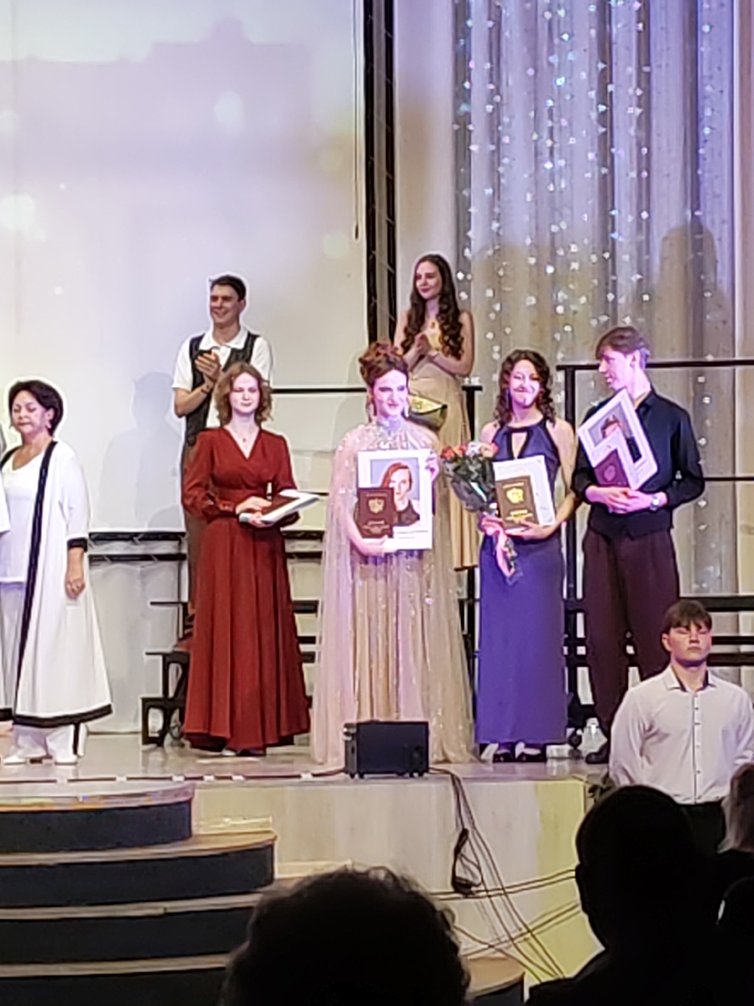 The image shows a group of people, likely graduates, on a stage. A man in a white shirt and vest is clapping. A woman in a red dress is holding a book and a certificate. Another woman in a purple dress is holding a bouquet of flowers and a certificate. A man in a black shirt and dark pants is holding a framed picture. In the background, there are curtains with sparkling decorations and a projection screen. The scene appears to be an awards ceremony or graduation event. The location is identified as Chaikovsky, Russia, although no specific landmarks are visible in the image to confirm this.