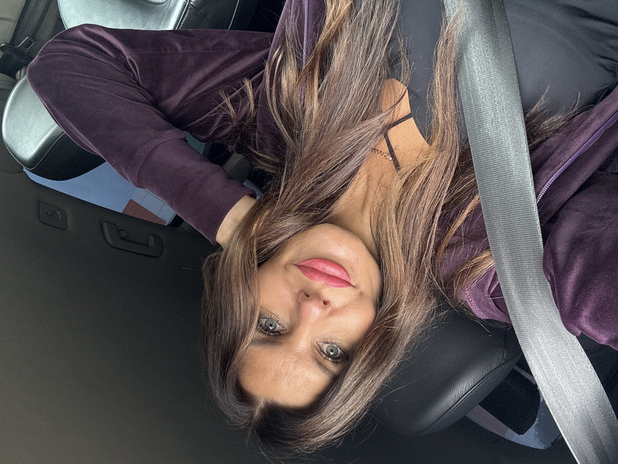 A woman with long, wavy brown hair is positioned inside a car, leaning her head back against the headrest. She is wearing a dark purple velour tracksuit and a black tank top with a thin gold necklace. A grey seatbelt is fastened across her chest. Her eyes are open, looking towards the camera, and she has bright pink lipstick on. The background shows the car's interior, including a black leather seat and part of a headrest. The lighting suggests it is daytime, and the image appears to be a selfie taken in the car. The scene does not contain any elements that suggest a specific location within the city of Кварса, Russia.