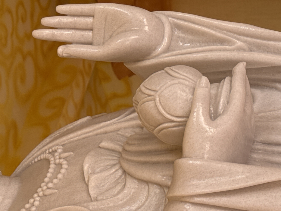 The image displays a close-up view of a light-colored stone statue, likely marble, located in Đăk Mar, Vietnam. The statue's upper torso and arms are depicted, featuring detailed drapery of robes. A double-stranded necklace with bead-like elements and pointed pendants is visible on the left side of the figure's chest. The figure's right hand is raised to shoulder height, fingers extended upwards with the palm facing forward. The left hand holds a closed lotus flower, cupped by the fingers. The background is a blurred, warm yellow-orange with subtle, indistinct patterns.