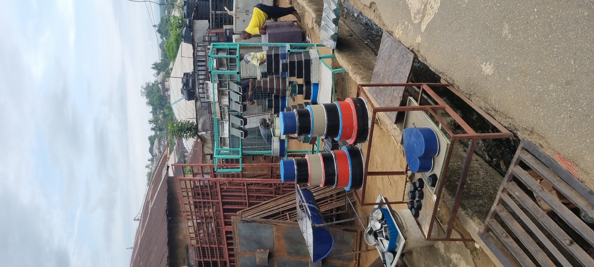 This media file depicts an outdoor metal fabrication or sales workshop, likely located in Calabar, Nigeria, during the daytime under an overcast sky.
The scene shows several individuals engaged in activities related to metalwork. In the background, a man in a yellow t-shirt and dark trousers is leaning over a large, black cylindrical object, possibly a tank. Near the center, another man wearing a plaid shirt is focused on arranging or working with smaller metal items on a display stand.
The workshop is filled with various metal products. Large black water tanks are visible in the background on the left. In the mid-ground, numerous light-colored metal sheets, possibly roofing or paneling, are neatly stacked on green metal racks. Dominating the foreground and center are several sturdy metal stands displaying colorful, circular metal objects in blue, black, red, beige, and silver, which could be industrial spools, decorative components, or covers. Below these, smaller blue heart-shaped or shield-shaped metal pieces and small black circular knobs or caps are arranged on separate display structures. A blue metal structure in the lower left foreground has the visible text "GE. ELECTRICAL". Various reddish-brown metal frames, rebar, and construction materials are also scattered around the site.
The workshop is set beside a paved road or path, with a drainage ditch covered by a metal grate running along its edge. The background features green tropical vegetation and residential buildings, all under a bright but cloudy sky. The overall impression is that of a bustling, functional outdoor commercial or industrial space.