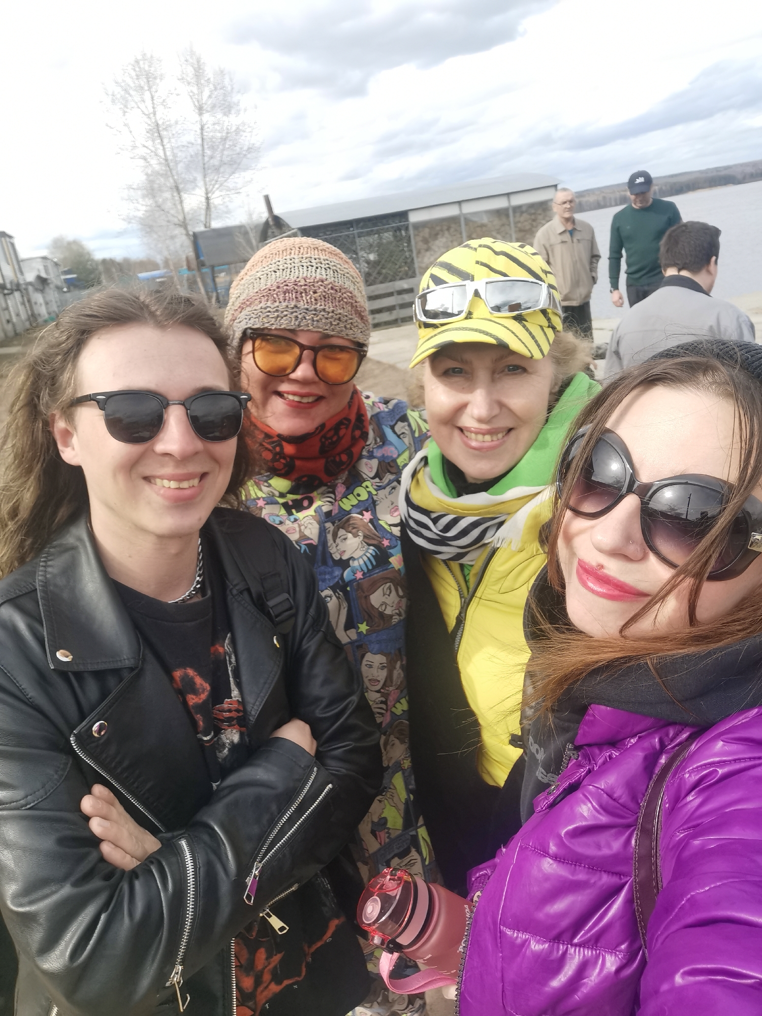 The image shows a group of four women taking a selfie outdoors. The woman on the left is wearing a black leather jacket, a black t-shirt, and sunglasses, with her arms crossed. Next to her is a woman in a multi-colored knit hat, orange-tinted glasses, and a jacket with cartoon faces printed on it. To their right is a woman with a yellow and black striped cap and a yellow jacket. The woman on the right is wearing large, dark sunglasses and a shiny purple jacket, and she is holding the camera.
In the background, there are three men standing near the edge of a body of water. There's also a structure resembling a boathouse and some trees in the distance. The sky is overcast. The location appears to be a waterfront area, possibly a park or recreational area.