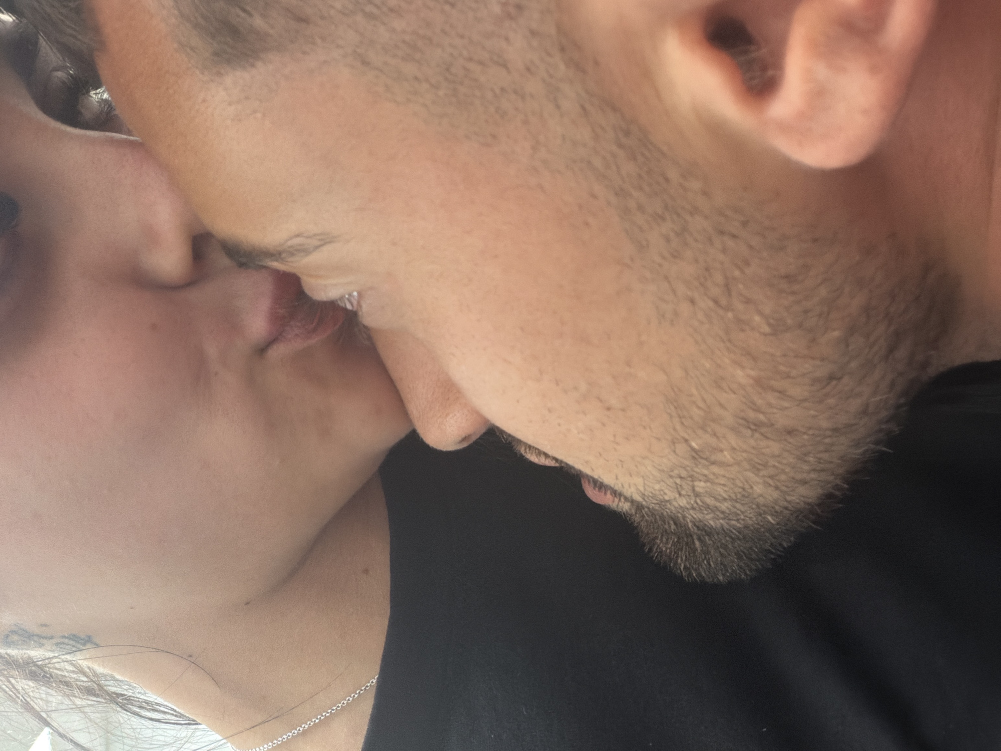 A close-up, low-angle shot captures a moment of intimacy between two individuals. The focus is on their faces, with one person's forehead gently touching the other's cheek as they share a kiss. The individual on the left, whose face is partially visible, has dark hair and is wearing a black top, with a silver chain necklace visible. The other individual, on the right, has short, dark hair and a light beard. Their skin tones appear to be similar. The lighting suggests an indoor setting, possibly a dimly lit room, with a soft glow illuminating their faces. No specific details about the location in Plovdiv, Bulgaria, are discernible from the image.