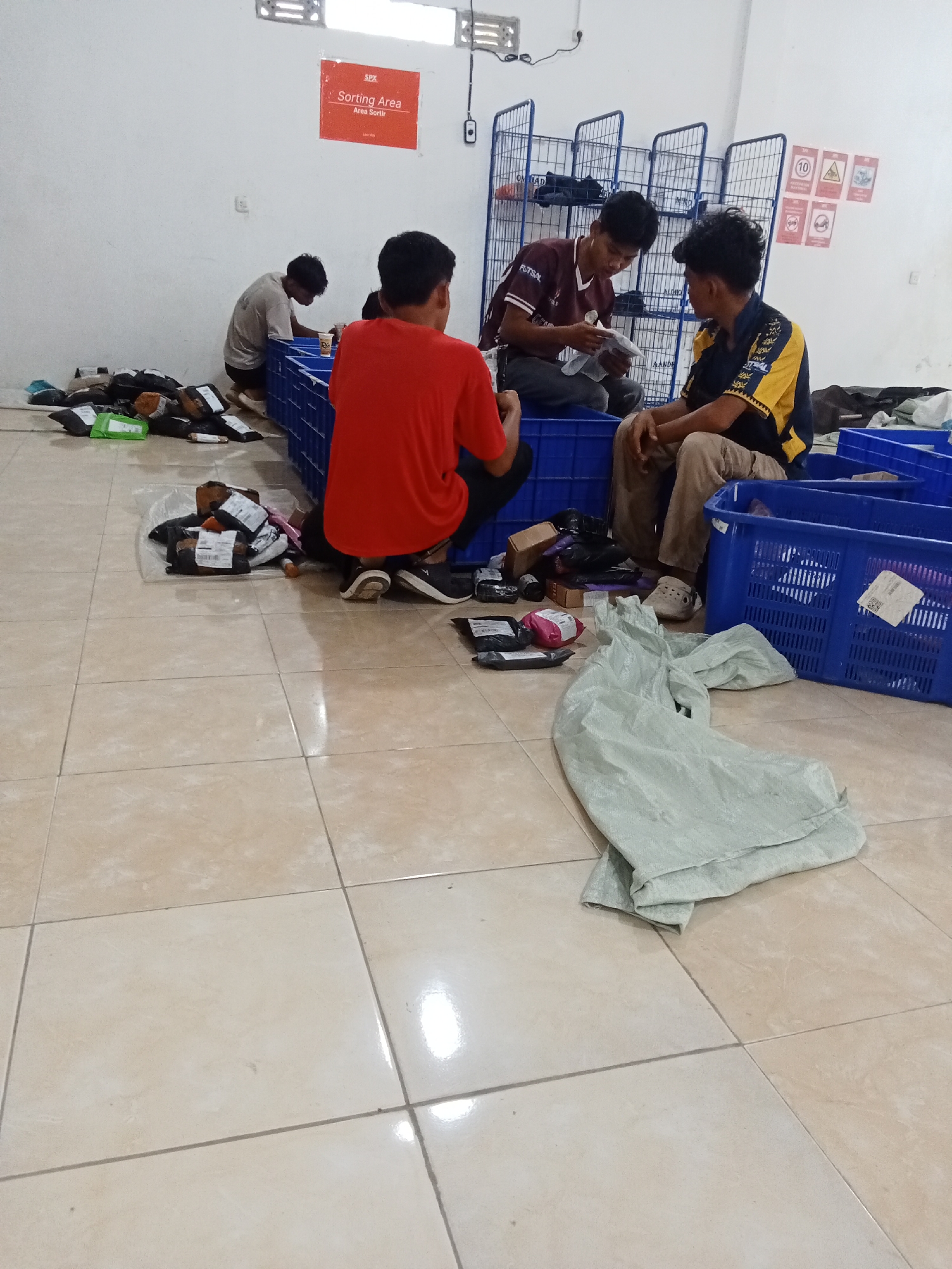 In Jirak, Indonesia, four young men are actively engaged in sorting and packaging items in what appears to be a warehouse or distribution center. The floor is tiled with light beige squares.
In the foreground, a pile of light green fabric lies on the floor. To the right of this fabric is a large blue plastic crate filled with items. To the left of the fabric, several packages are scattered on the floor, some wrapped in brown paper and others in clear plastic.
In the mid-ground, three of the men are interacting with these packages and crates. One man, wearing a red t-shirt and dark pants, is kneeling and appears to be working with a package. Next to him, another man in a dark t-shirt with the word "FUTSAL" on it is sitting and holding a white item, possibly a label or document. To the right of these two men, a third man, wearing a yellow and blue collared shirt, is seated and also handling some papers.
In the background, a fourth man, dressed in a light-colored t-shirt and dark pants, is kneeling and working with packages near a white wall. Above this area, a red sign with white text reads "SPX Sorting Area Area Sortir." Behind the sorting area, several tall blue metal shelving units are visible, holding various items. On the wall behind the shelves, several red and white warning signs are affixed. The environment is well-lit with fluorescent lighting from above.