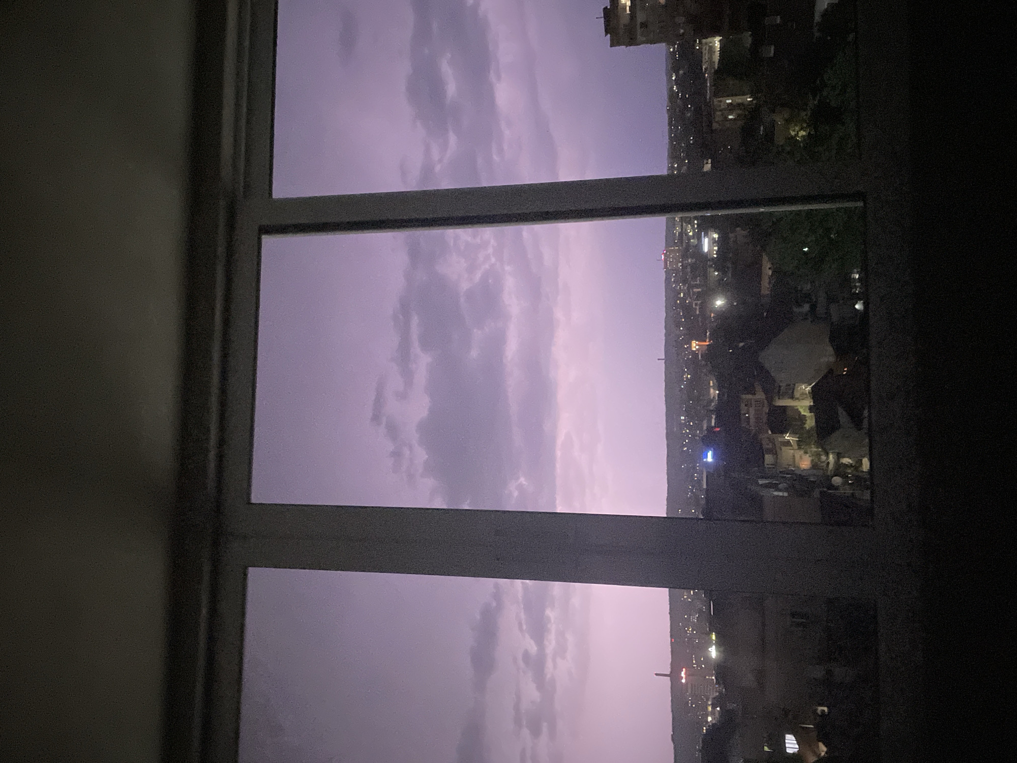 A view from a window looking out over a city at dusk. The sky is a pale purple with clouds, and the city lights are visible in the distance. The window frame is white. The location is unknown.
