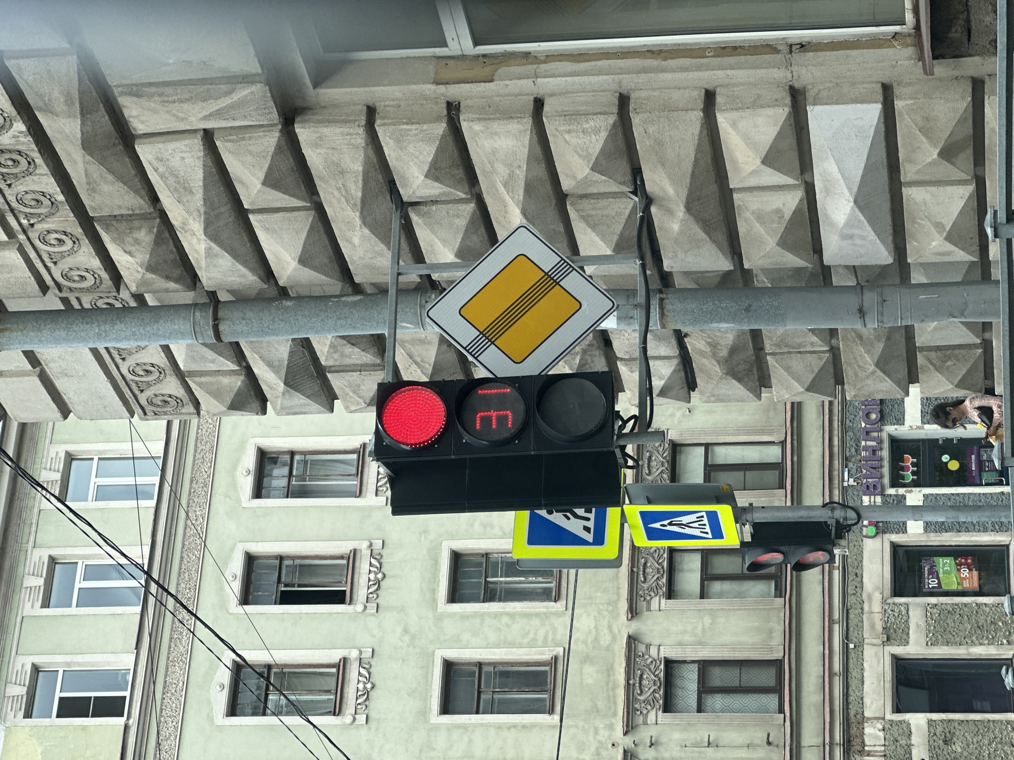 The image shows a street scene in Saint Petersburg, Russia, featuring a traffic light with a red signal and a digital countdown timer displaying "31". Adjacent to the traffic light are two pedestrian crossing signs. The building on the right has a textured facade with geometric patterns, while the building on the left is a pale green color with multiple windows. A person wearing a light-colored top and sunglasses is visible in the lower right corner, looking at their phone. The environment appears to be an urban setting.