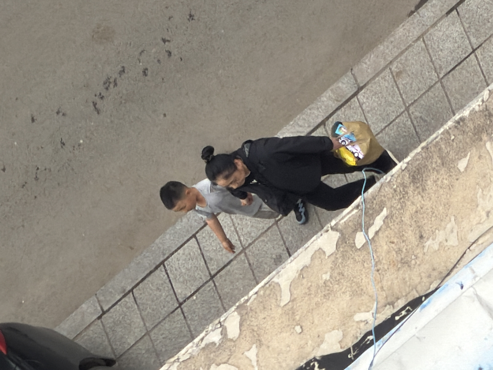 An overhead photograph captures two individuals, an adult and a child, walking along a paved outdoor area in Tongjiang, China. The scene is viewed from an elevated perspective, looking down at the ground and subjects.
The adult, likely female given her hairstyle (hair tied in a bun), is dressed in a black jacket and black pants, with blue and black sneakers. She is carrying a brown paper bag in her left hand, which contains several colorful items. One visible item appears to be a small package with cartoon-like images or a logo, and another is a bright yellow object, possibly a snack or small toy.
Beside her, a child, likely male, wears a light grey short-sleeved t-shirt and light-colored pants. His head is partially visible, with short dark hair. Both individuals are in motion, walking towards the upper left of the frame.
The ground beneath them is composed of light grey, square-patterned tiles or paving stones. To the right of the walking path is a low wall or ledge, appearing aged with patches of peeling lighter-colored material over a darker base. A thin, light blue string or wire hangs down near this wall. In the bottom left corner, part of a black vehicle, possibly a car, with a red detail is visible. The lighting is even, suggesting an overcast day or indirect sunlight. No specific text is clearly legible on any objects or surfaces. No strong emotions are discernible from the distant view of the subjects.