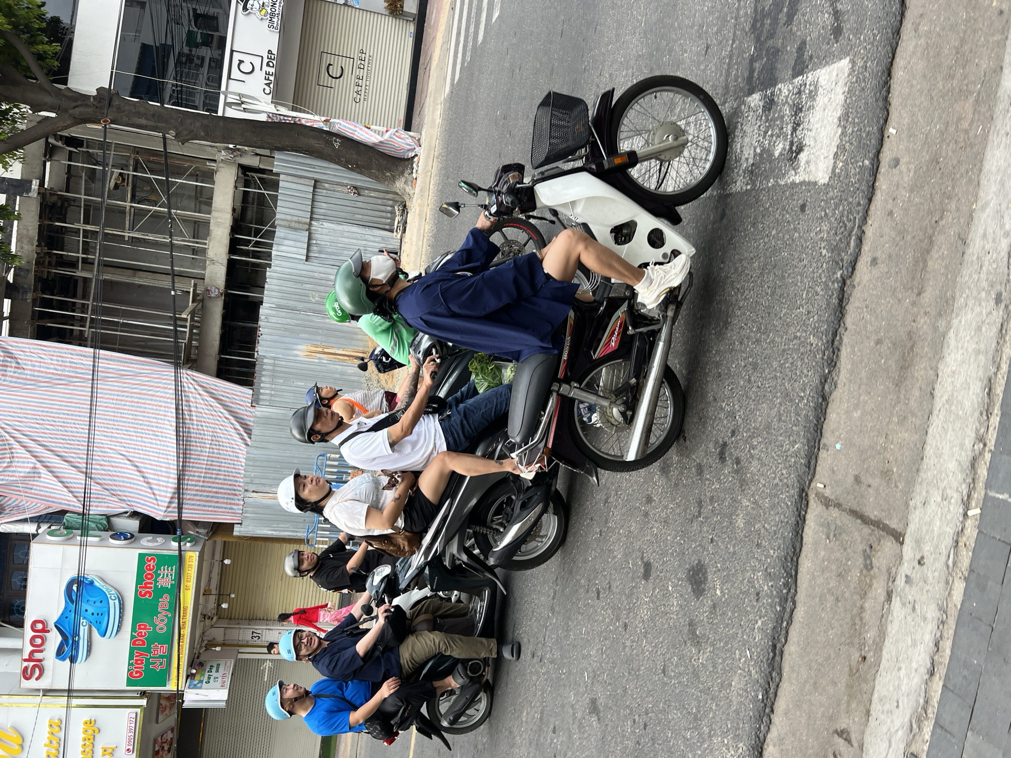 The image depicts a street scene in Nha Trang, Vietnam. Several individuals are riding on motorcycles. In the foreground, a person wearing a green helmet and a blue jacket is riding a white and black motorcycle. Behind them, a woman with a white helmet and a white shirt is seated on a motorcycle, holding a small dog. Further back, a man in a dark shirt and khaki pants is on a motorcycle, followed by a man in a blue shirt. In the background, there are buildings with shops and signs, including one for "Shop" with a picture of blue Crocs shoes, and another for "Cafe Dep." Construction scaffolding and corrugated metal sheeting are visible on one of the buildings. The street is asphalt, with a concrete curb and sidewalk. Power lines are strung across the top of the image. The overall atmosphere suggests a bustling urban environment.