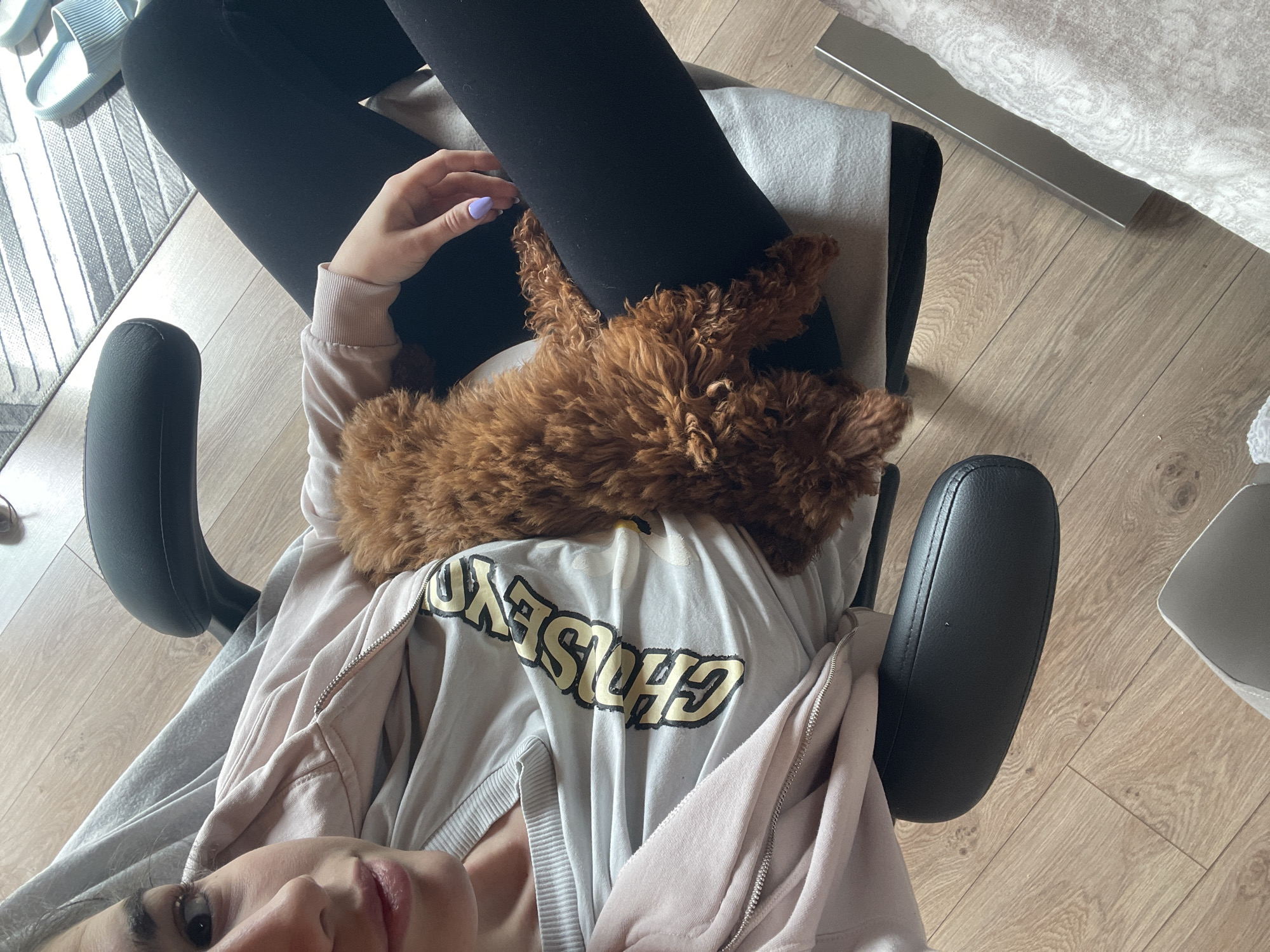 A person is sitting in a black office chair with armrests. The person is wearing black leggings, a light gray T-shirt with "CHLSSYDN" printed on it, and a light pink zip-up hoodie. A brown poodle-type dog is lying on the person's chest and lap. The location appears to be inside a room with light wood flooring. Blue sandals are visible on a striped rug to the left. Part of a bed with a patterned comforter is visible in the upper right corner. The photo appears to be taken from the person's perspective. There is no definitive evidence in the image to confirm the location is Sofia, Bulgaria.