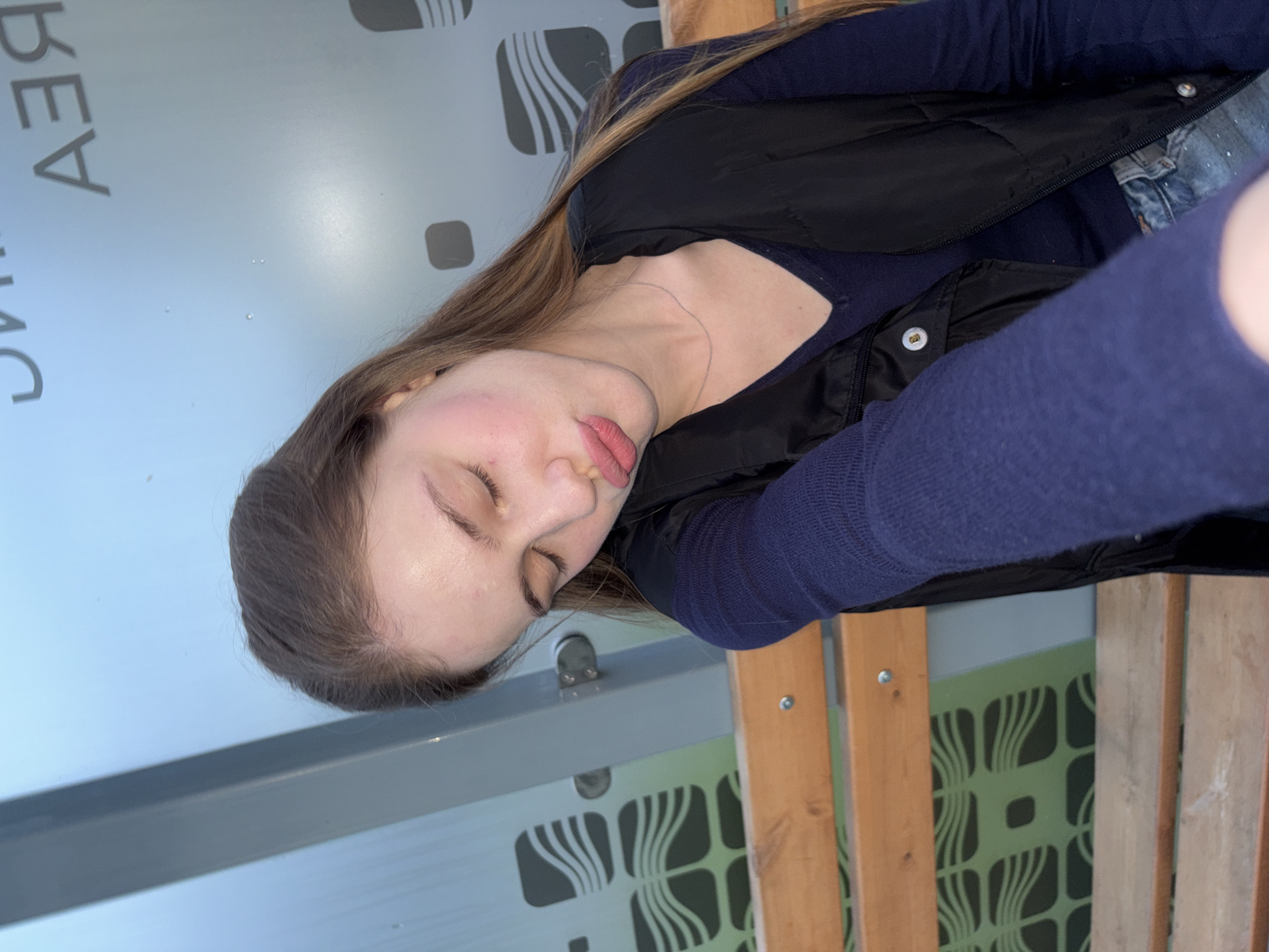 The image shows a young woman with her eyes closed and head tilted to the side. She is wearing a dark blue long-sleeved shirt and a black vest. She appears to be sitting on a wooden bench in an outdoor setting. Behind her, there is a glass panel with a geometric pattern and some text, partially obscured. The text visible appears to be in Cyrillic script. The lighting suggests it is either early morning or late afternoon. The location is identified as the city of Arfa, Russia.