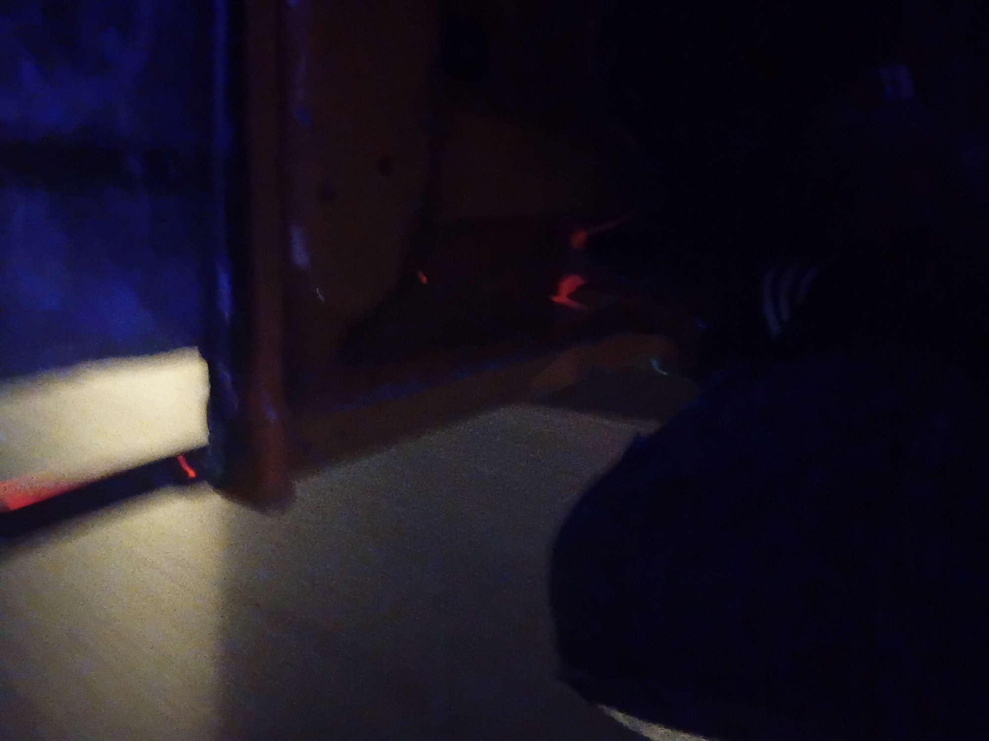 The image depicts a dimly lit interior, possibly a room or a vehicle, with an orange surface in the foreground that appears to be a table or a seat. The lighting is dramatic, with streaks of red and blue light casting long shadows. In the lower part of the image, a person's legs and lap are visible, covered in dark clothing. The environment seems to be in the city of Jalingo, Nigeria, though specific details of the location are not discernible from the image. The scene is somber and mysterious, with the low light and the obscured details contributing to an atmosphere of unease.