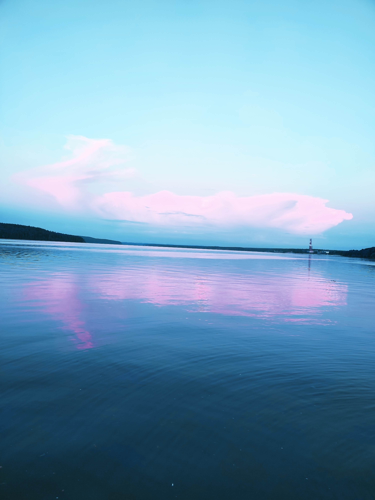 A vertical, wide-angle shot captures a serene body of water with a pale blue and pink sky overhead. A large, fluffy cloud formation, tinged with pink, dominates the upper half of the sky, stretching horizontally across the frame. The water's surface is calm, with gentle ripples reflecting the sky's colors, creating a pinkish hue that contrasts with the deeper blue of the water. A dark, tree-lined shoreline runs along the horizon in the background, with a white and red striped lighthouse visible in the distance on the right. The overall impression is one of peaceful twilight or dawn.