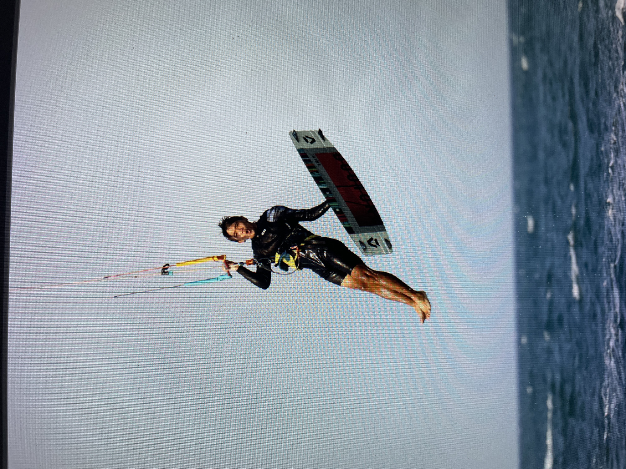 A man is kiteboarding in the city of Bacong, Philippines. He is airborne, holding onto a yellow and blue kite control bar and connected to a kiteboard. The sea is to his right.