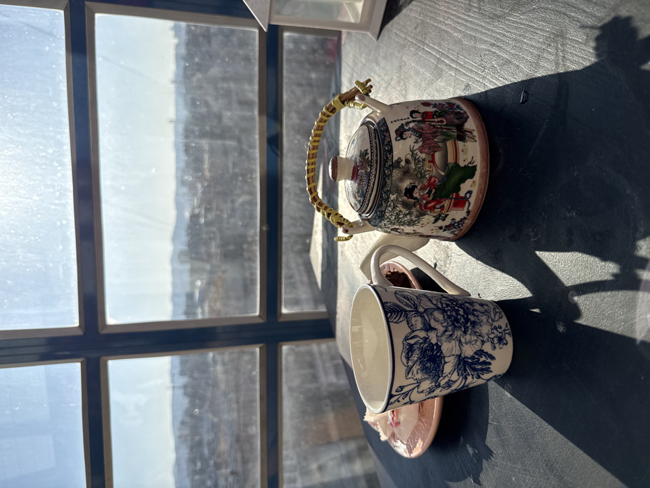 An elevated interior scene in Krasnoyarsk, Russia, viewed on a sunny day. The foreground features a dark, reflective surface, likely a table, upon which several objects are arranged. To the left, a white ceramic mug with a blue floral pattern rests. To its immediate left, a small, light pink plate holds a white pastry with red elements. To the right of the mug, a decorative ceramic teapot with an Asian-inspired design, depicting figures in traditional attire and floral motifs, sits prominently. The teapot has a braided yellow and brown handle. Further to the right, partially visible, is a small white decorative lantern.
Strong, direct sunlight streams from the background, casting distinct shadows of the mug and teapot onto the table surface towards the lower left. The background is dominated by a large, multi-paned window, through which a panoramic view of an urban landscape is visible. Buildings and structures populate the mid-ground, while distant hills or mountains are discernible on the horizon under a clear, bright sky. Lens flare and light haze are present in the upper sections of the window, indicating intense sunlight. No individuals are present in the frame.