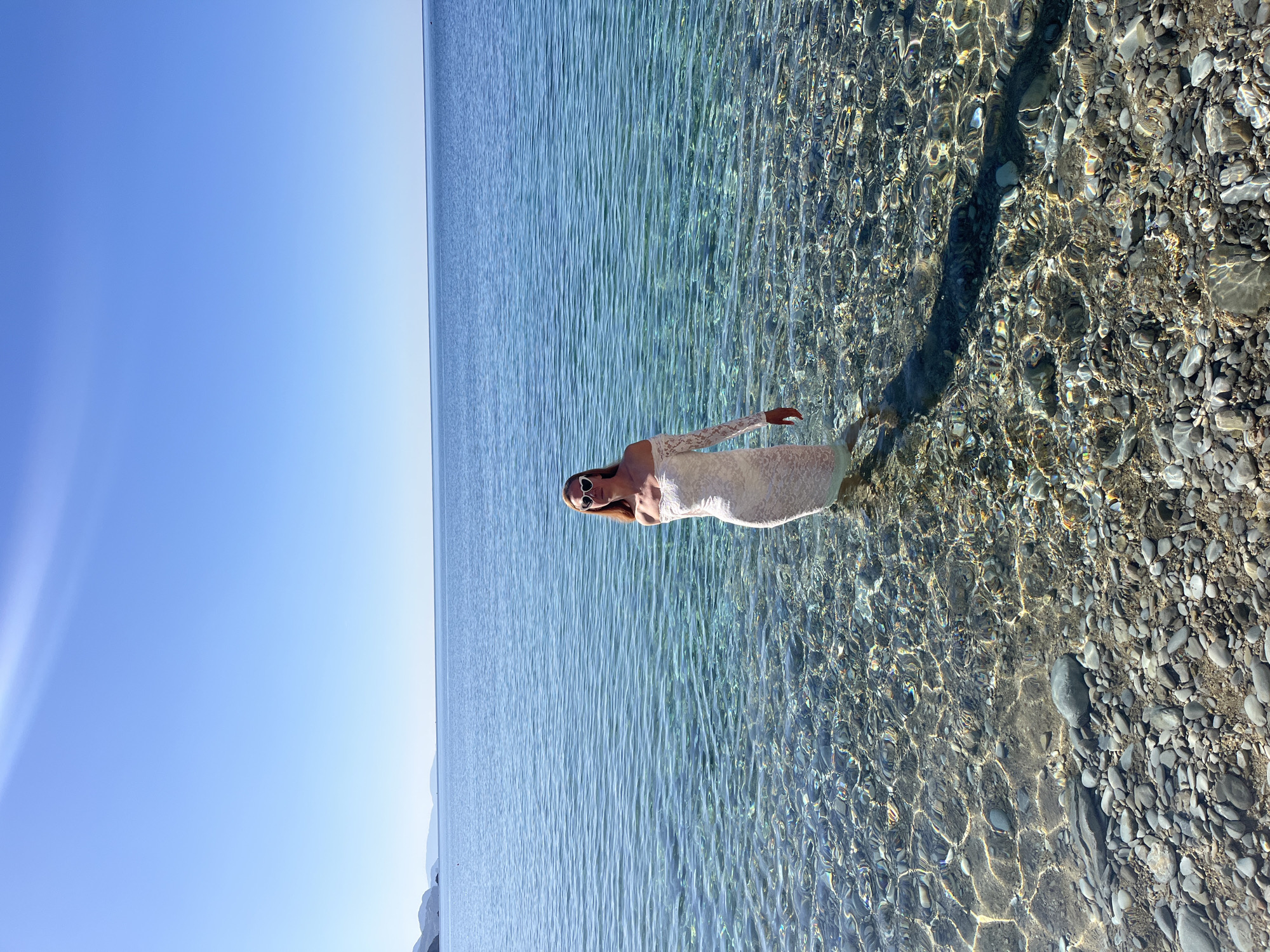 A woman stands in the shallow, clear water of a beach near Budva, Montenegro. She is wearing a light-colored dress with lace sleeves and sunglasses. The water is clear enough to see the pebbles and stones on the seabed. The sky is clear blue. The Adriatic Sea stretches to the horizon. The image appears to be taken in the daytime, with the sun positioned to the right, casting a shadow of the woman in the water.