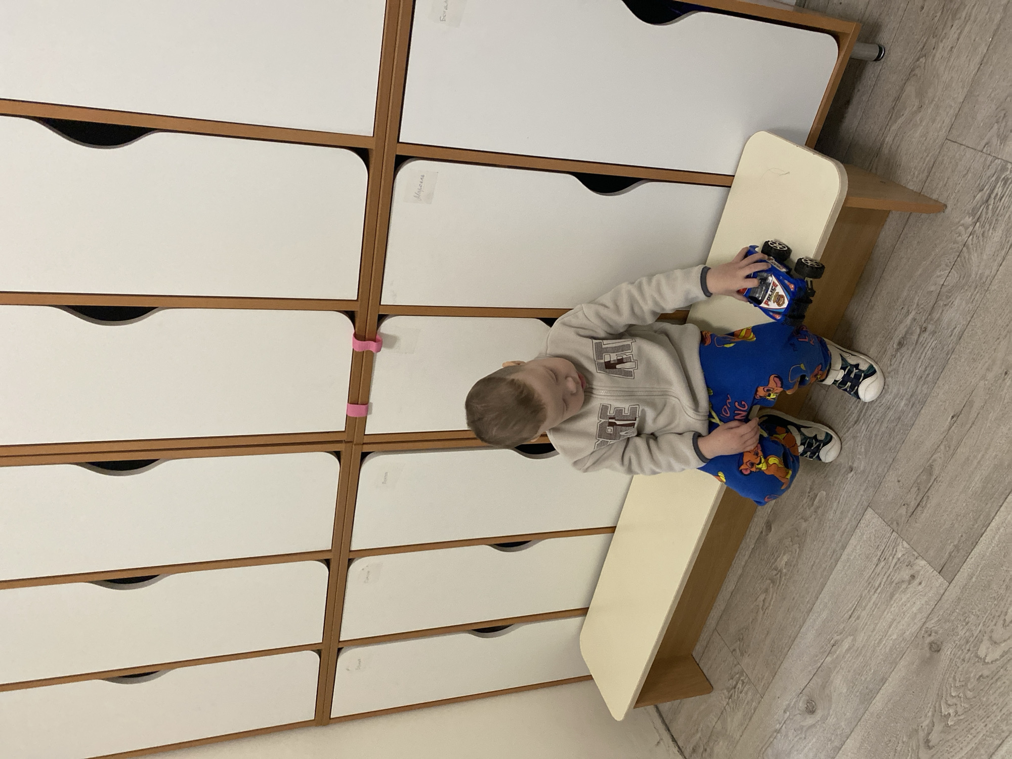 A child sits on a small bench in what appears to be a child care setting or preschool. The child is wearing a light gray sweatshirt and blue pants with cartoon images, along with black and white sneakers. The child is holding a blue toy car. Behind the child is a wall of white storage lockers with wooden frames. The room has light-colored wood-patterned flooring. There is no definitive evidence in the image to confirm this scene is in Sochi, Russia.