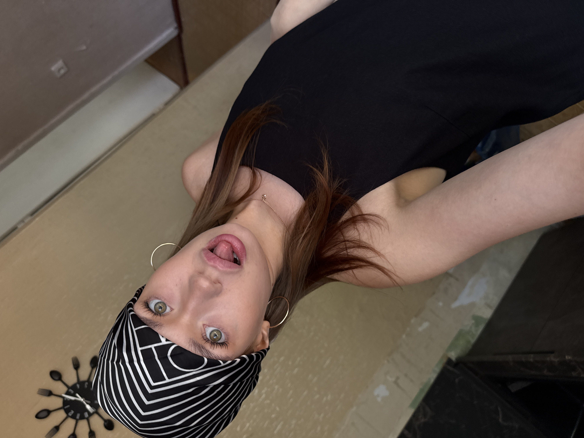 The image shows a young woman lying down, with her head tilted back and her tongue sticking out. She is wearing a black bandana with a white striped pattern, a black top, and a necklace. She also has hoop earrings. Her eyes are wide open and looking at the camera. The background is blurry but appears to be an indoor setting with a wooden floor and a wall. There is a clock on the wall behind her, which is unusual as it has forks and spoons instead of numbers. The lighting suggests it is daytime, but there are no specific clues about the weather. The woman's expression is playful and provocative. The image was taken in Chaikovsky, Russia.