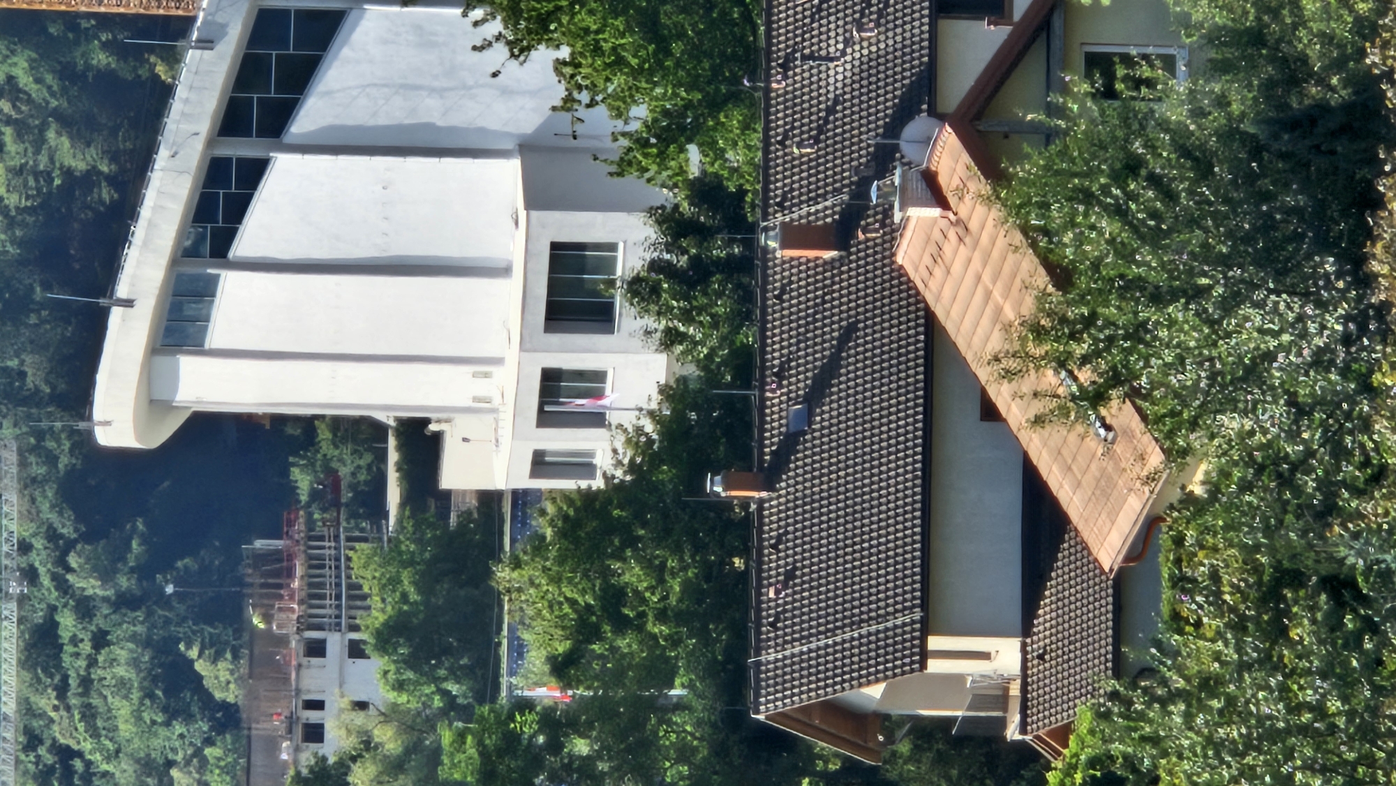 The image shows a view of buildings and lush green trees, likely on the outskirts of a city, given the presence of construction in the background. A large, modern white building with a curved roof and large windows dominates the upper right portion of the frame. Below it, several residential buildings with tiled roofs are visible, some appearing older and in need of repair, while others are newer. A construction site is partially visible in the upper left, with scaffolding and what appears to be a worker in a yellow vest. The trees are dense and varied in species, suggesting a temperate climate. No specific landmarks or features that definitively place the image in Prešov, Slovakia, are present. The scene appears to be a daytime shot, with bright sunlight casting shadows.