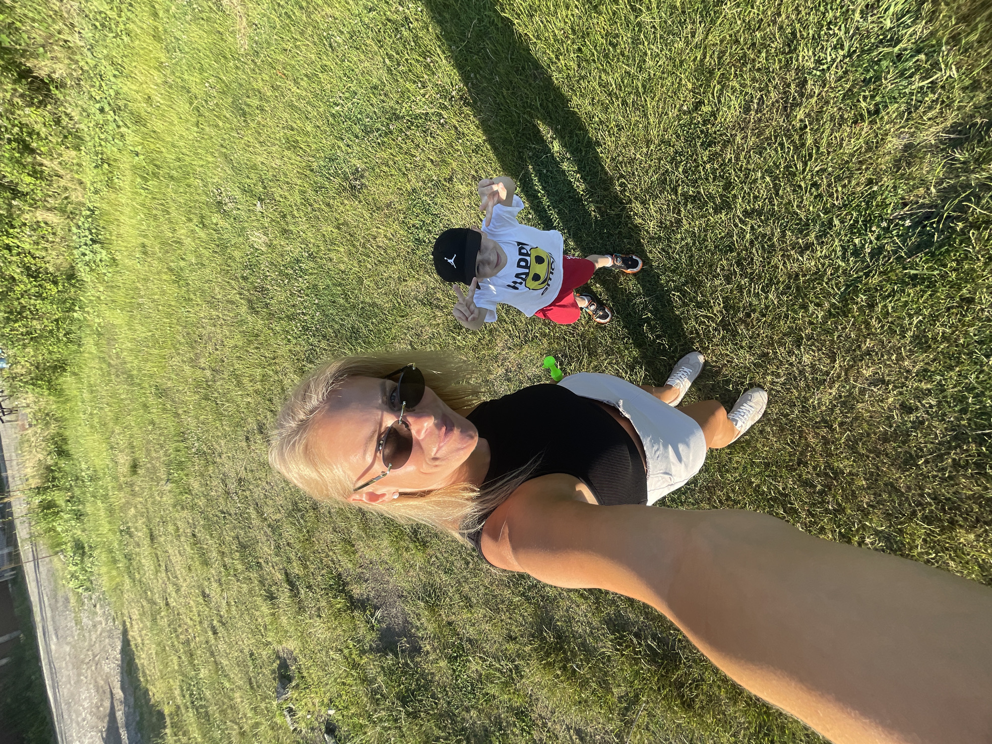 This is a selfie taken outdoors in a grassy area, likely a park or yard, given the presence of green grass and a shadow cast by trees. The image features two people: a woman lying on the grass and a young boy standing.
The woman is in the foreground, lying on her back with her head tilted slightly. She has blonde hair, is wearing sunglasses, a black top, and white shorts. Her right arm is extended, holding the camera. She appears relaxed and happy, with a slight smile.
The boy is in the background, standing and looking up at the camera. He is wearing a white t-shirt with a smiley face graphic and the word "HAPPY" printed on it, red shorts, a black baseball cap, and sneakers. He is making a peace sign with his fingers on both hands. He appears energetic and joyful.
The scene is bathed in natural sunlight, suggesting it is daytime. The long shadows indicate it could be late afternoon or early morning. The overall atmosphere is one of leisure and enjoyment.
The text visible on the boy's shirt reads "HAPPY" with a smiley face graphic. There is also a Jordan logo on his cap.
The location context is Baranovka, Russia.