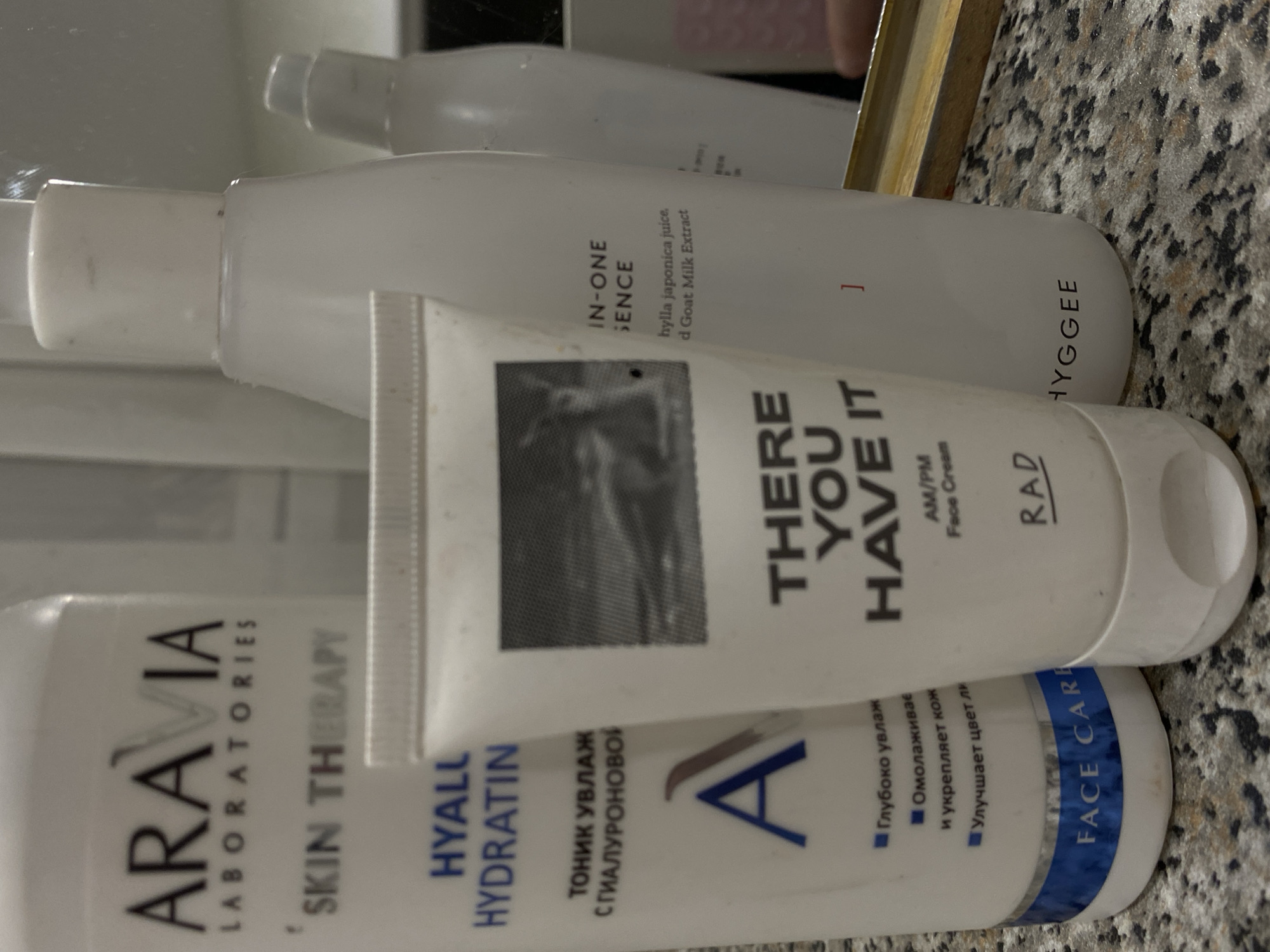 The image shows three white cosmetic bottles with text on a speckled surface that appears to be a countertop. The topmost bottle is partially visible and seems to be a cosmetic cream. The middle bottle has "THERE YOU HAVE IT AM/PM Face Cream" and "RAD" printed on it. The bottom bottle reads "ARAVIA LABORATORIES SKIN THERAPY HYALL HYDRATIN" with Russian text and the brand logo. It is not possible to determine the location in Kazan, Russia, from the provided image.