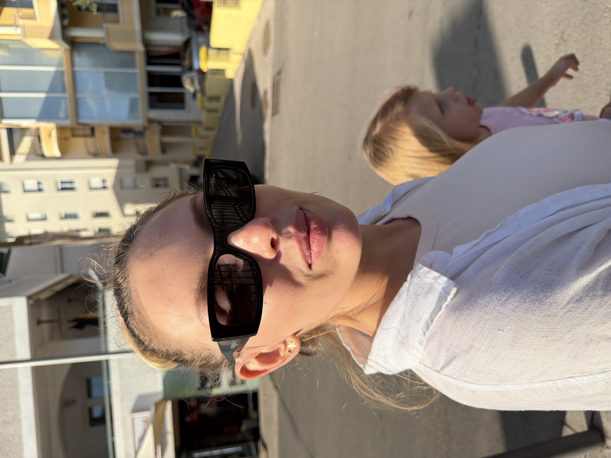 A woman wearing black sunglasses and a white shirt stands in the foreground. Behind her, a young child with blonde hair is visible. The background shows buildings and a street with yellow elements. The scene appears to be on a sunny day. The location is identified as Budva, Montenegro.