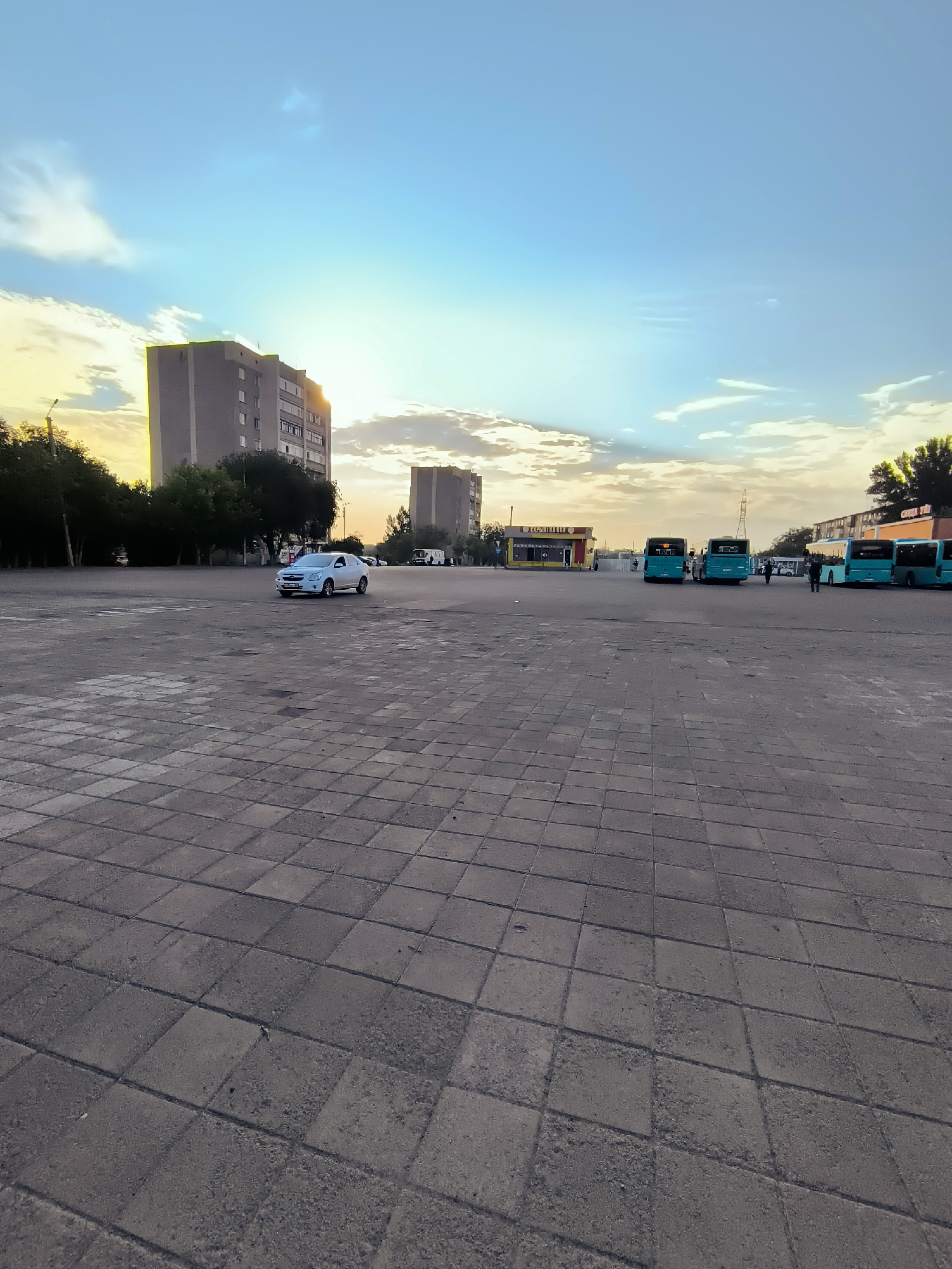 The image depicts an open paved area, likely a plaza or parking lot, in front of several buildings. The paving stones are laid in a grid pattern. In the mid-ground, a light-colored sedan is driving across the paved area. Further back, a row of teal-colored buses is parked. Behind the buses, low-rise buildings with storefronts are visible, one of which has yellow and red signage. On the left side of the image, taller residential buildings are present, some partially obscured by trees. The sky is a gradient of blue, with scattered clouds illuminated by the setting or rising sun, casting a warm light on the scene. The overall impression is a moderately urban environment during twilight. This scene is consistent with the area near the Qarağandı city administration in Kazakhstan, known for its open spaces, multi-story buildings, and public transportation hubs.
