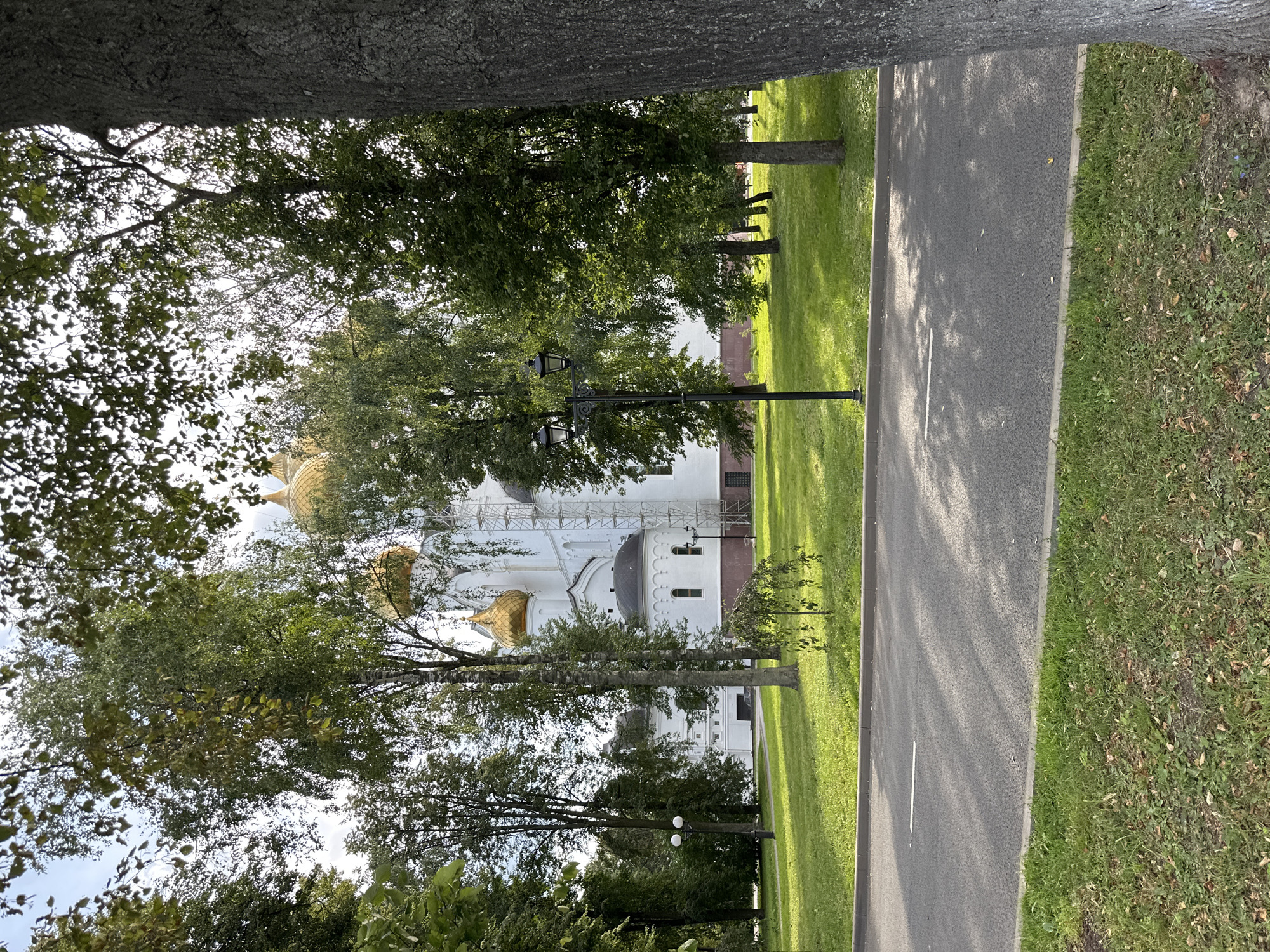 The image depicts a view of the Yaroslavl Assumption Cathedral in Rusanovo, Russia, on a sunny day. The cathedral, a white building with golden domes, is partially obscured by lush green trees. A paved road runs in the foreground, separated from a well-maintained lawn by a curb. A lamppost stands on the lawn. The overall scene is tranquil, with dappled sunlight filtering through the leaves of the trees, casting shadows on the road and grass.
