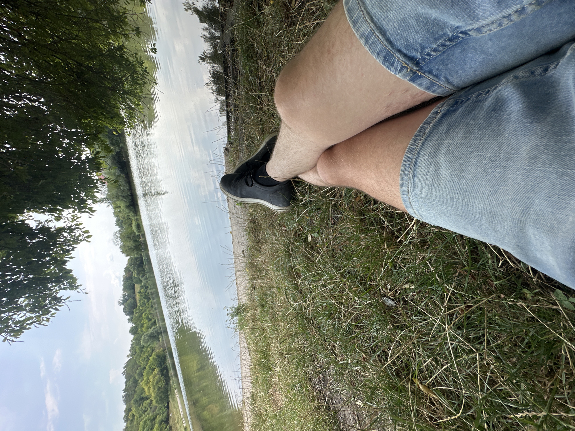 A low-angle, medium shot captures a person's legs, clad in light-wash denim shorts and dark sneakers, resting on a grassy bank beside a calm lake. The water reflects the sky and the lush green trees that line the opposite shore, creating a serene natural landscape. Overhead, the branches of a large tree with green leaves extend into the frame, casting dappled shadows. The scene appears to be set in the vicinity of Makiv, Ukraine, given the surrounding natural environment of a lake and dense vegetation. The weather seems fair, with a bright sky and scattered clouds, suggesting a pleasant day outdoors.